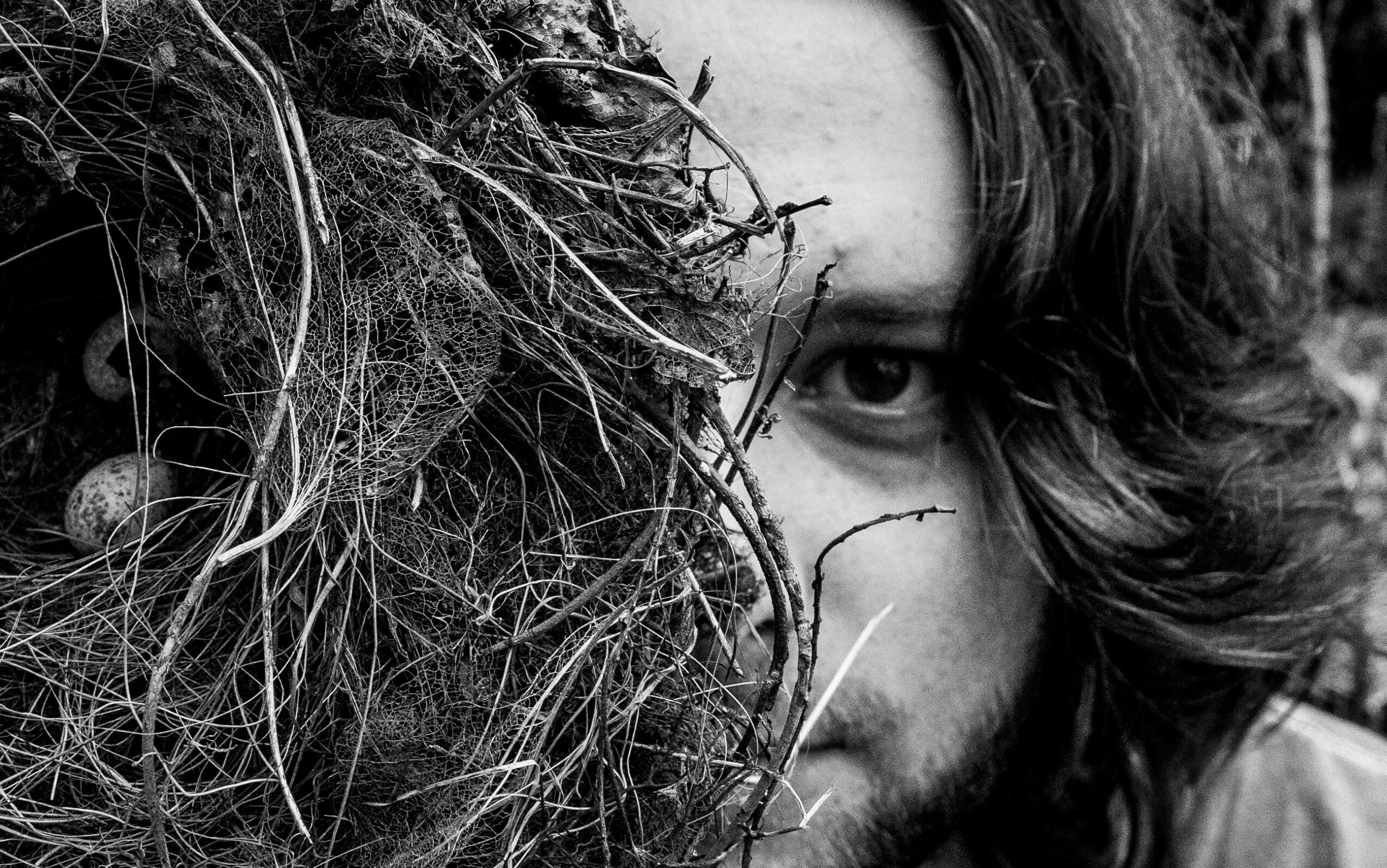 Person with long hair partially hidden behind a large nest or tangle of twigs and branches, with one eye visible.