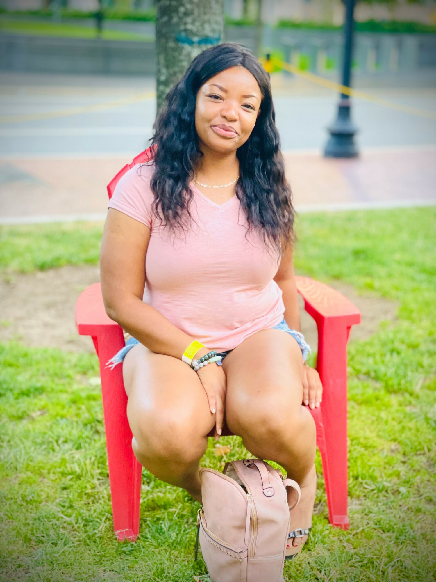 Young woman sitting on a red chair outdoors, smiling, wearing a pink shirt and denim shorts, with a park background.