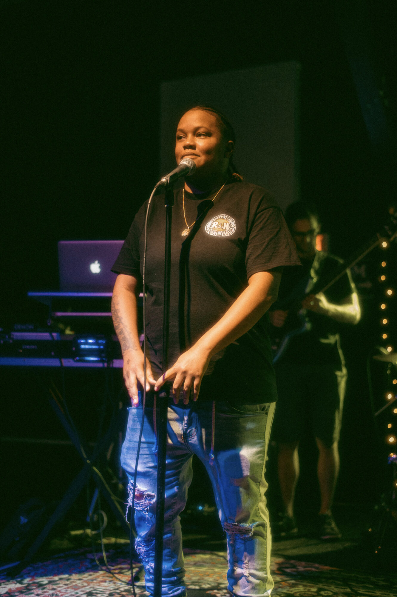 Woman standing on stage with microphone, wearing a black shirt and jeans, with a person in the background, stage lights visible.