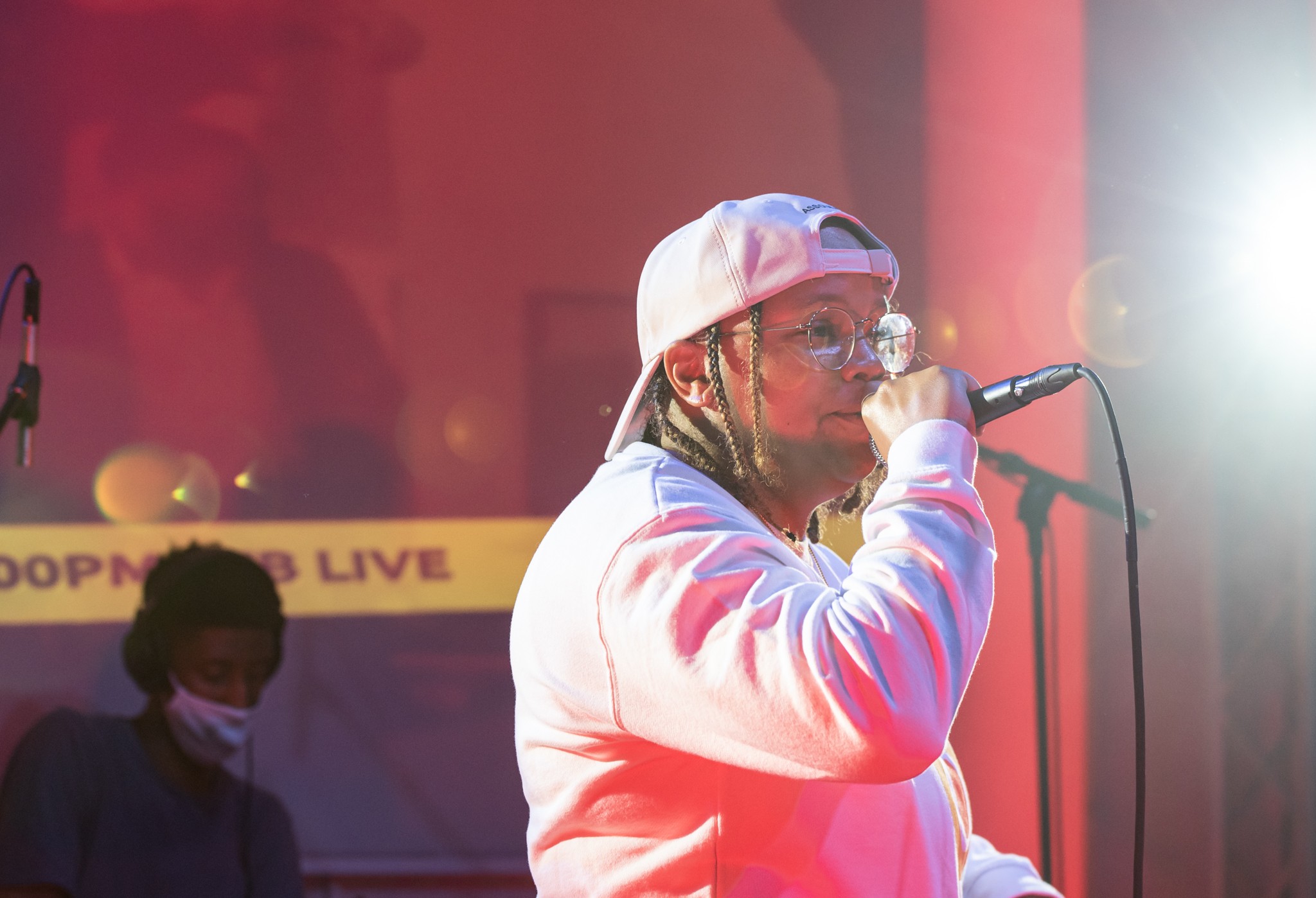 Person wearing a white hoodie and cap, holding a microphone, performing on stage with bright lighting.
