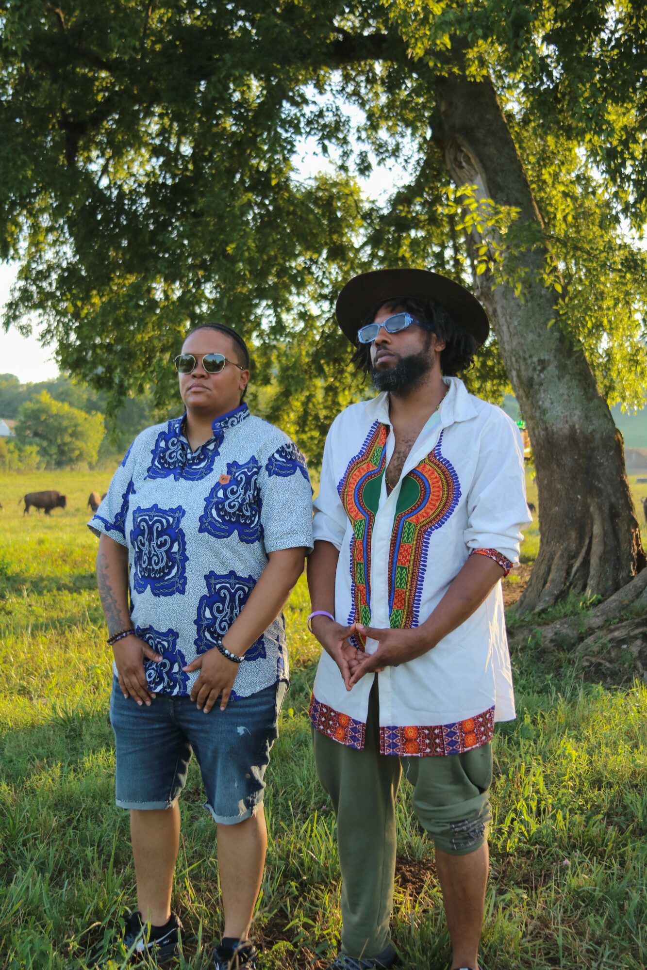 Two people standing outdoors under a large tree, one wearing a blue patterned shirt and sunglasses, the other in a colorful shirt and wide-brimmed hat, with a grassy field and cows in the background.