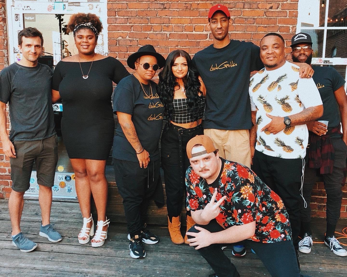 Group of eight people standing outdoors in front of a brick wall, smiling and posing for a photo.