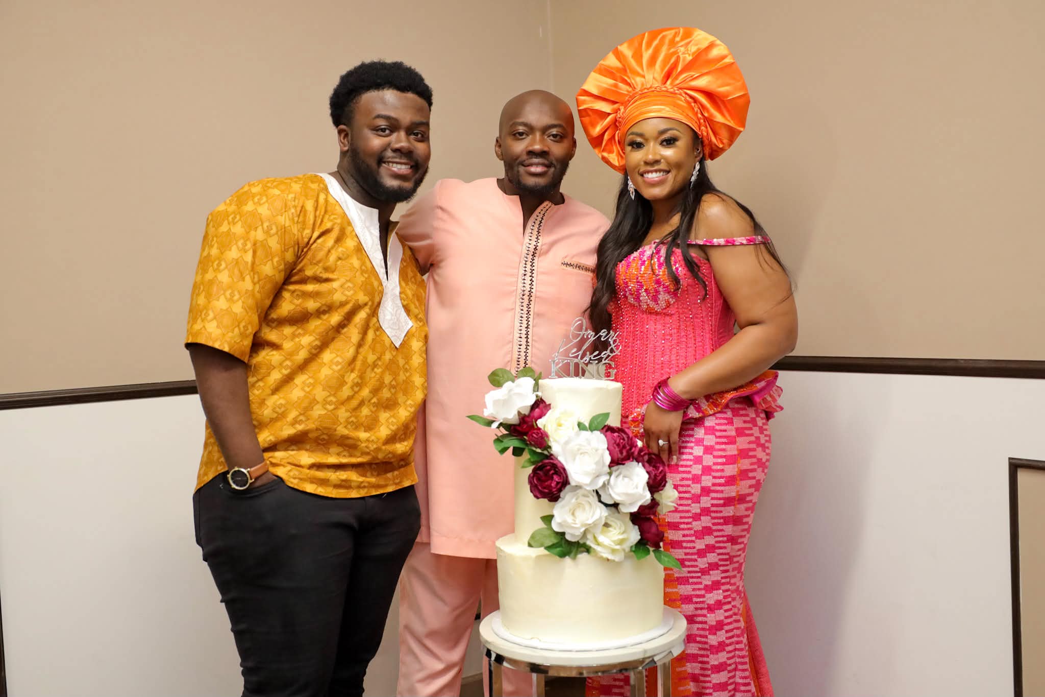 Three people standing together, smiling, with a decorated cake in front of them, against a plain background.