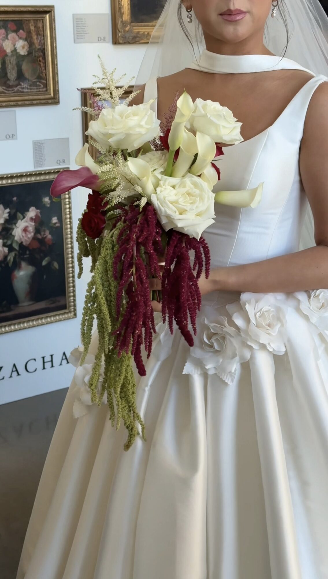 Woman in white dress holding a bouquet of white and burgundy flowers, with framed paintings in the background.