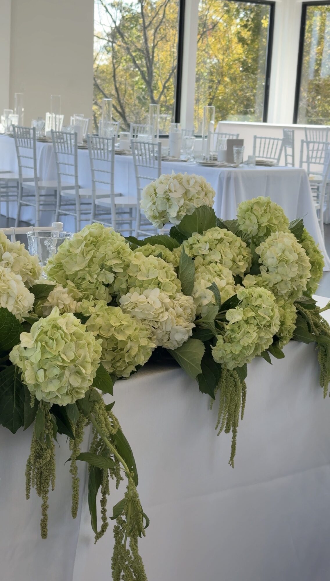 White hydrangea flowers arranged on a table in a bright, elegant event space with large windows and white chairs.