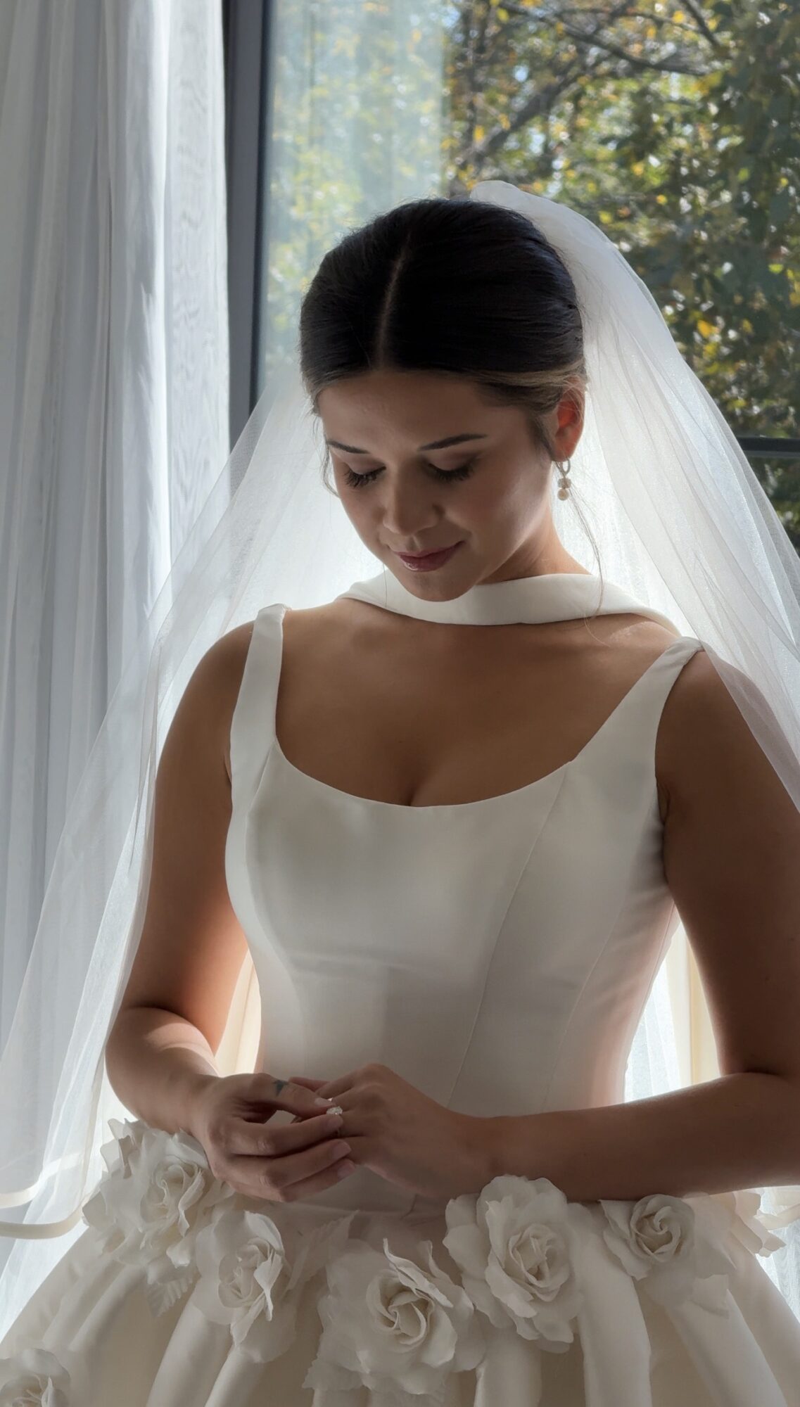 Bride in a white dress with floral details, standing near a window with sunlight, looking down with hands clasped.