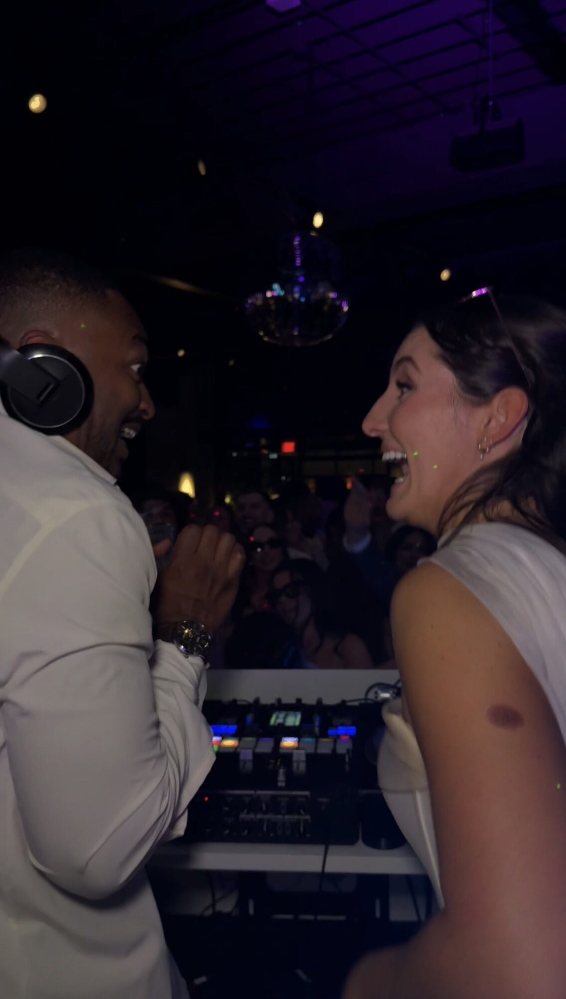 Two women smiling and talking at a party with a DJ setup, dim lighting, and a disco ball overhead.