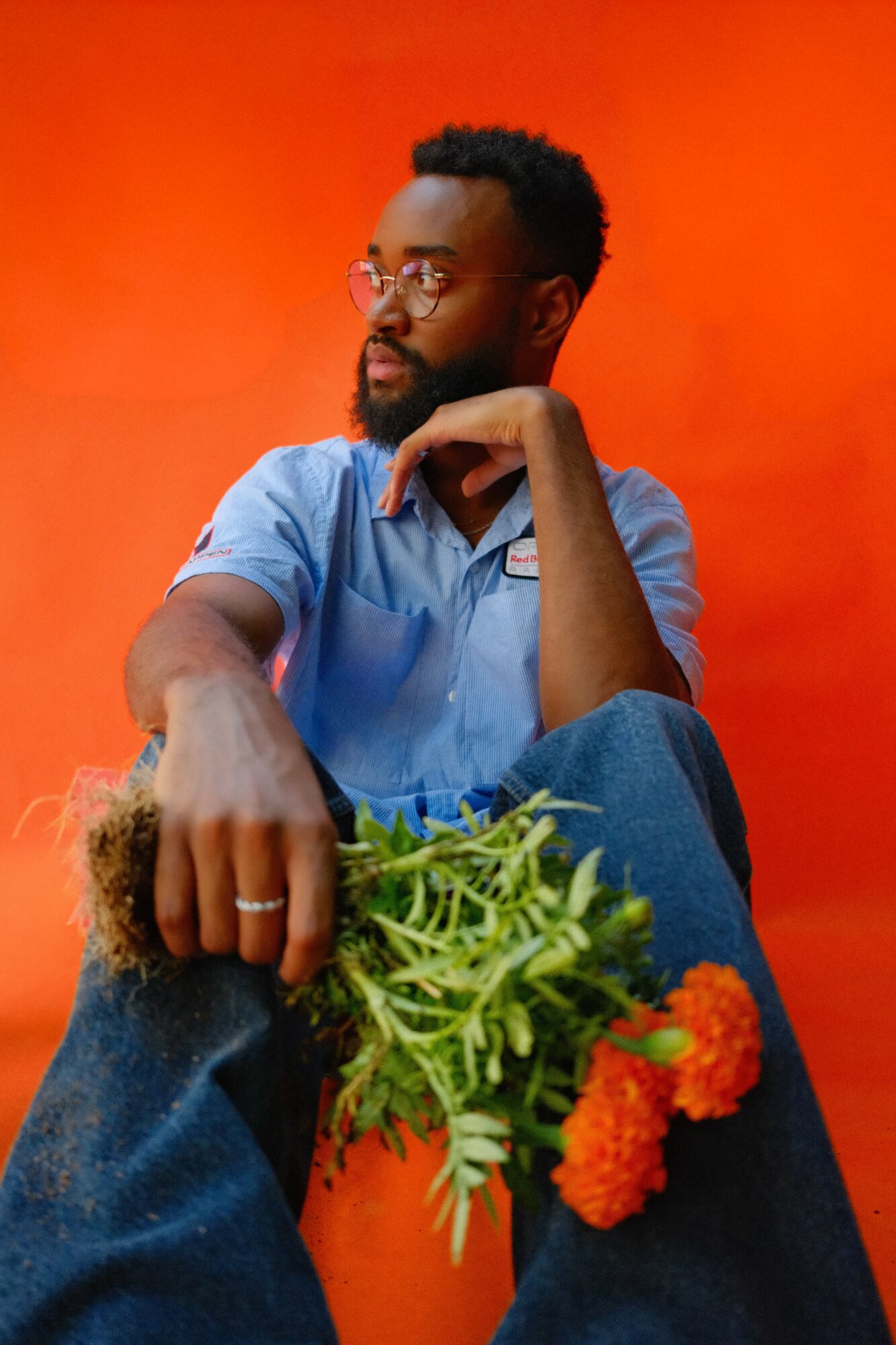 Man with glasses and beard sitting against orange background, holding a bunch of green plants and orange flowers, looking to the side.