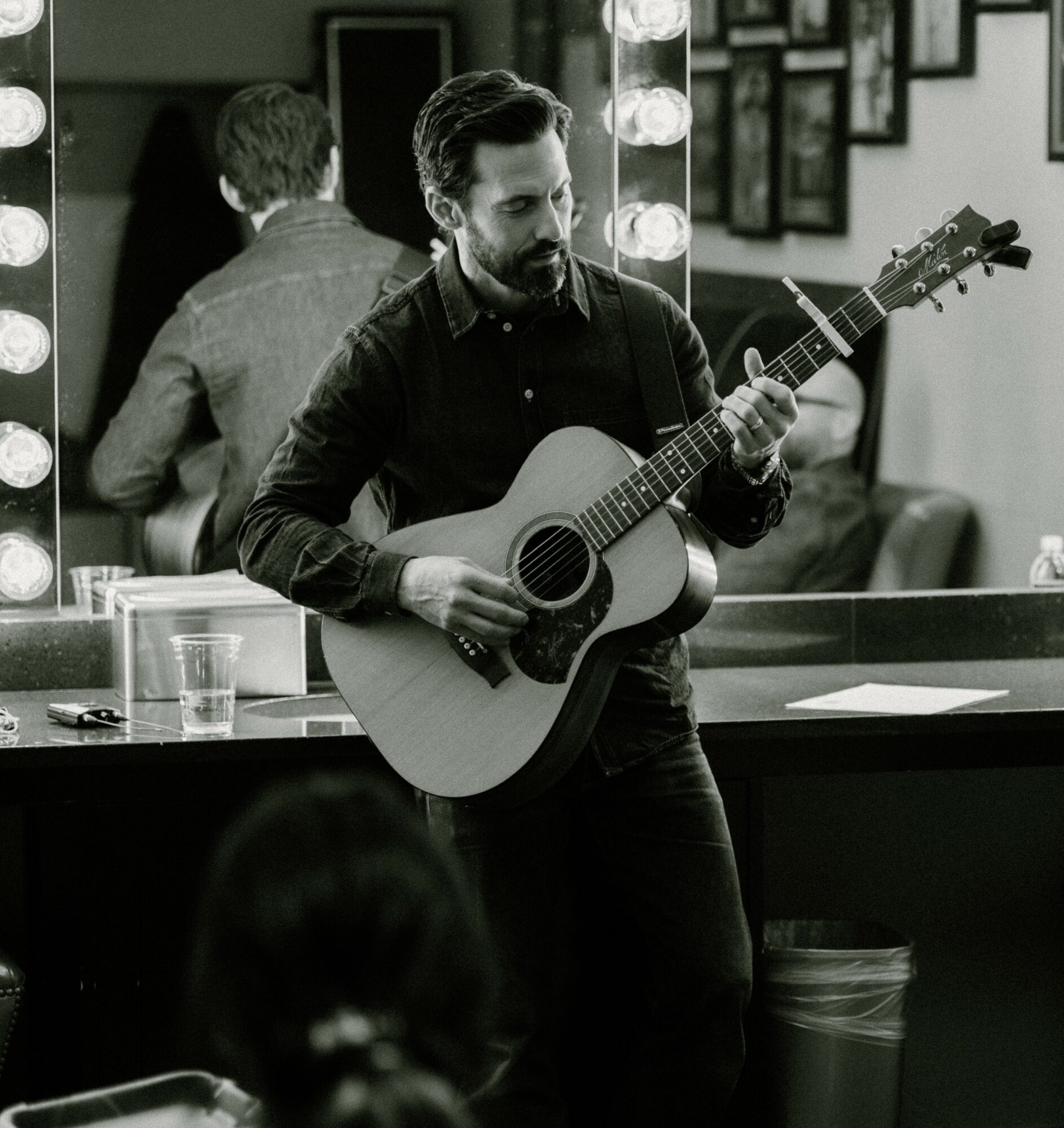 Man playing guitar in a room with mirrors and framed pictures, with a person in the foreground and a bar counter nearby.
