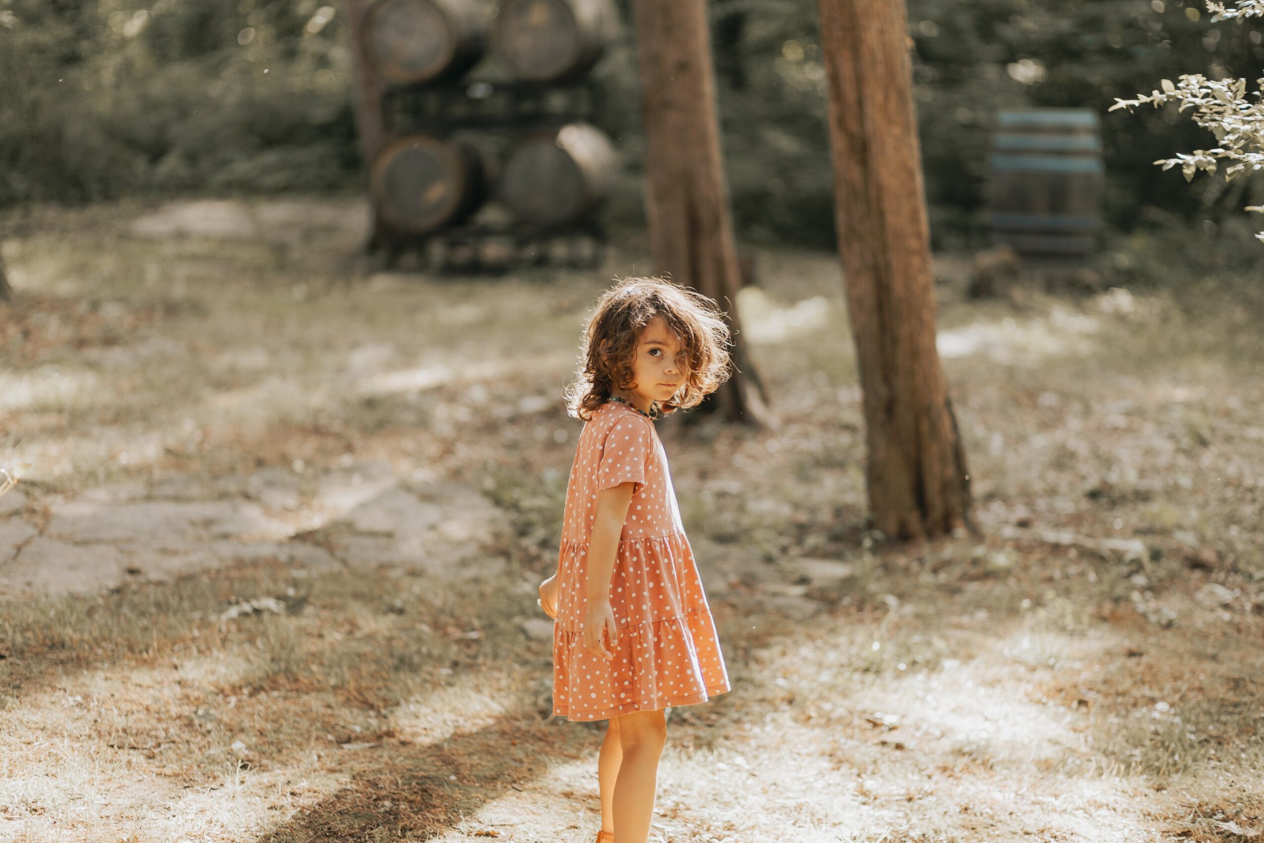 Young girl with curly hair in an orange dress standing outdoors on a sunny day.