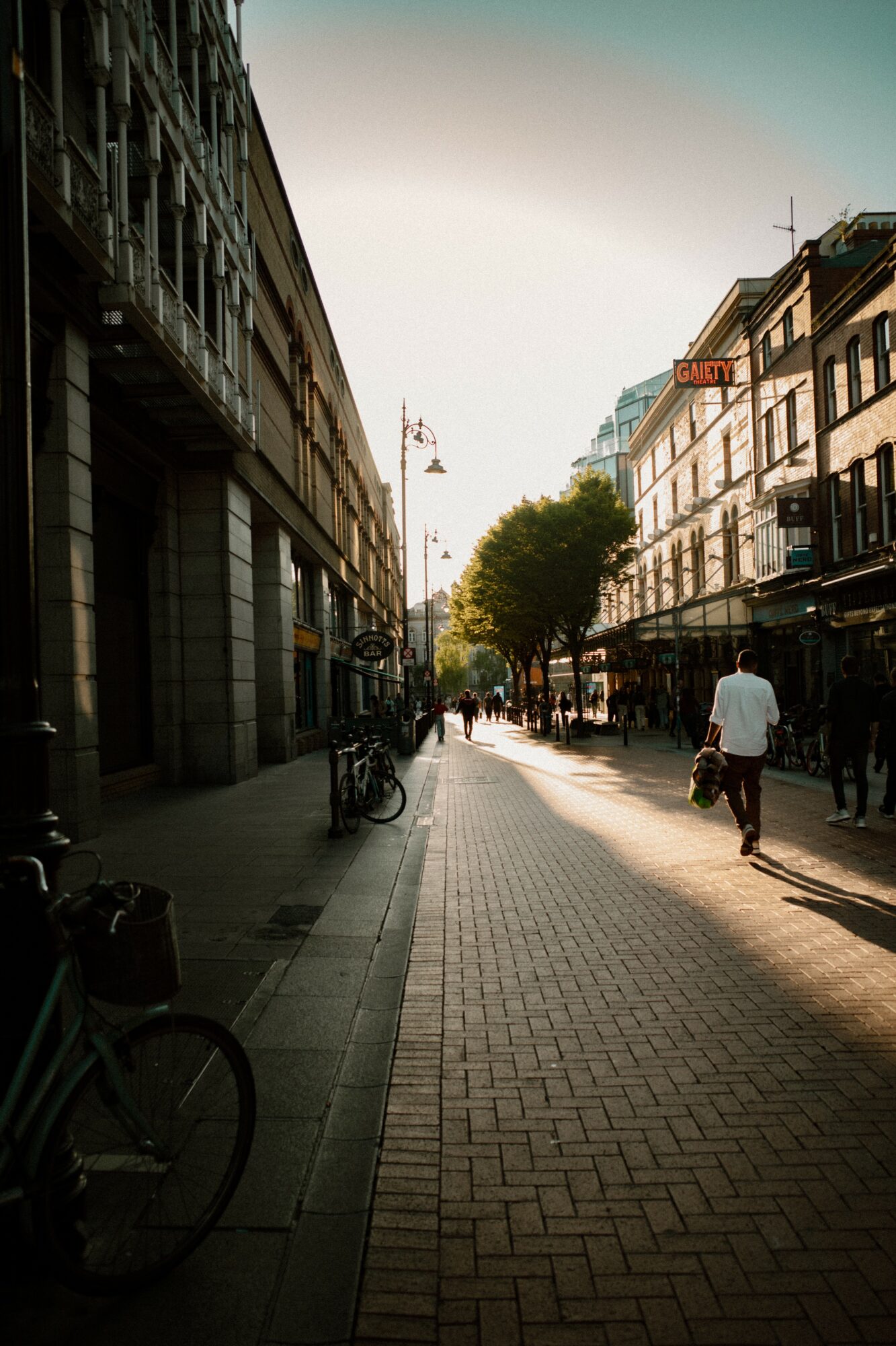 Pedestrian street with buildings, trees, and people walking, sunlight casting shadows, bicycles parked along the sidewalk.