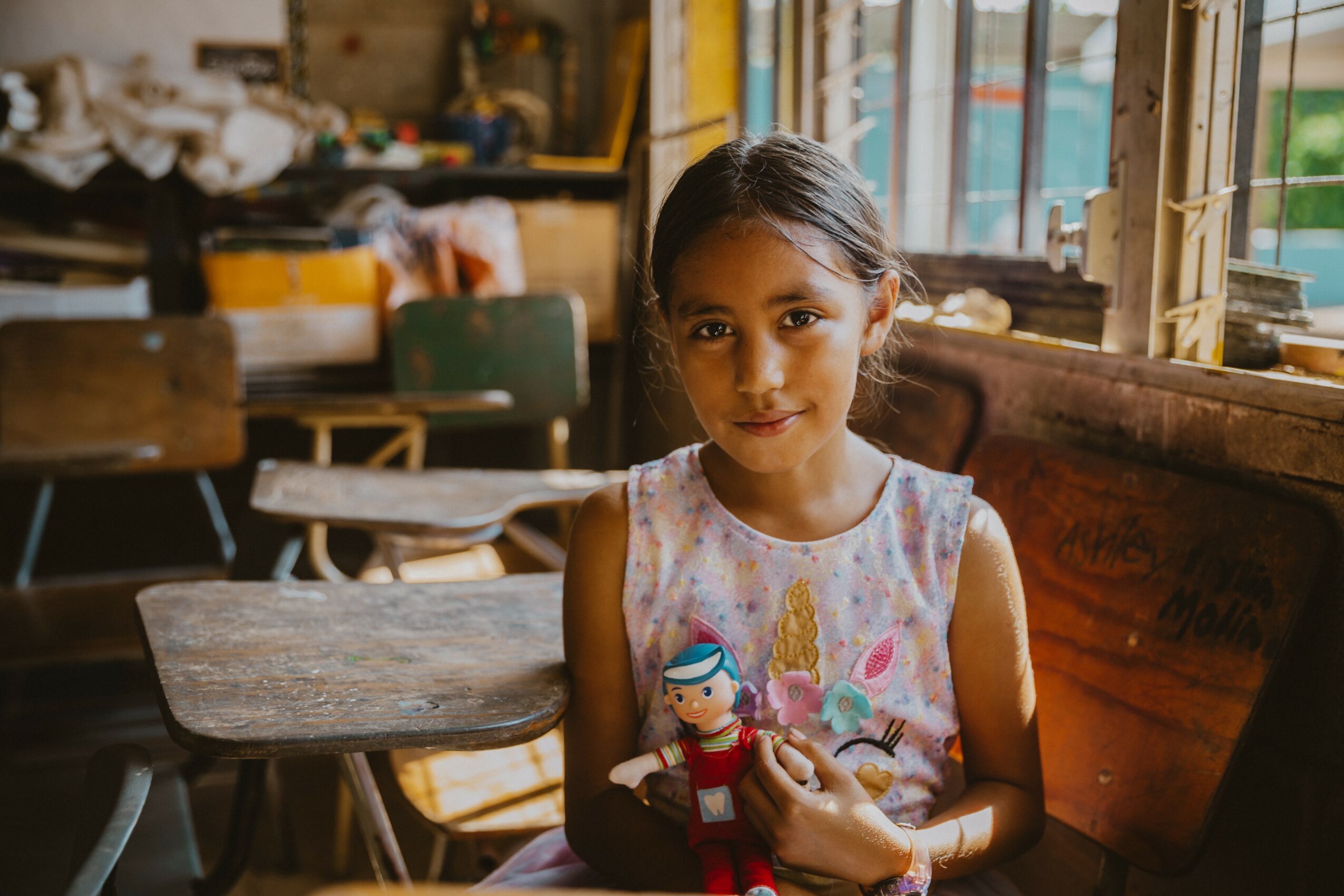 Young girl sitting in a classroom holding a doll, with desks and windows in the background.