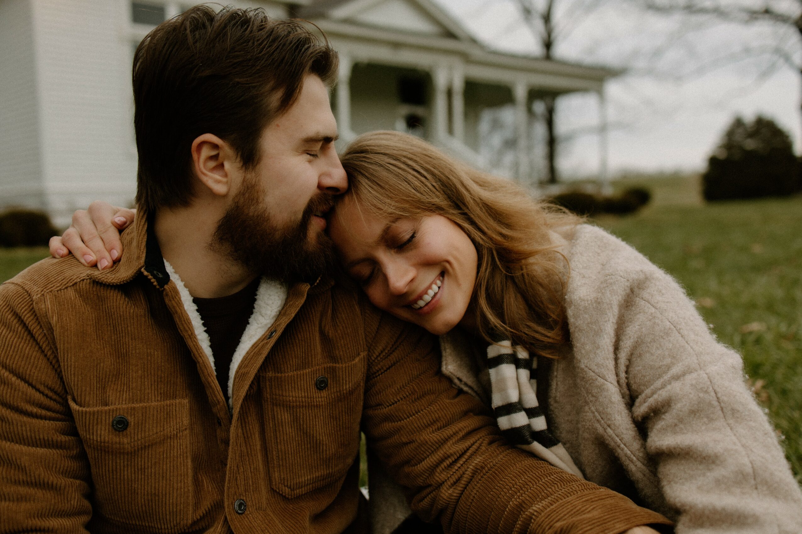 A smiling woman leans her head on a man's shoulder outdoors, both appear happy, with a house and trees in background.