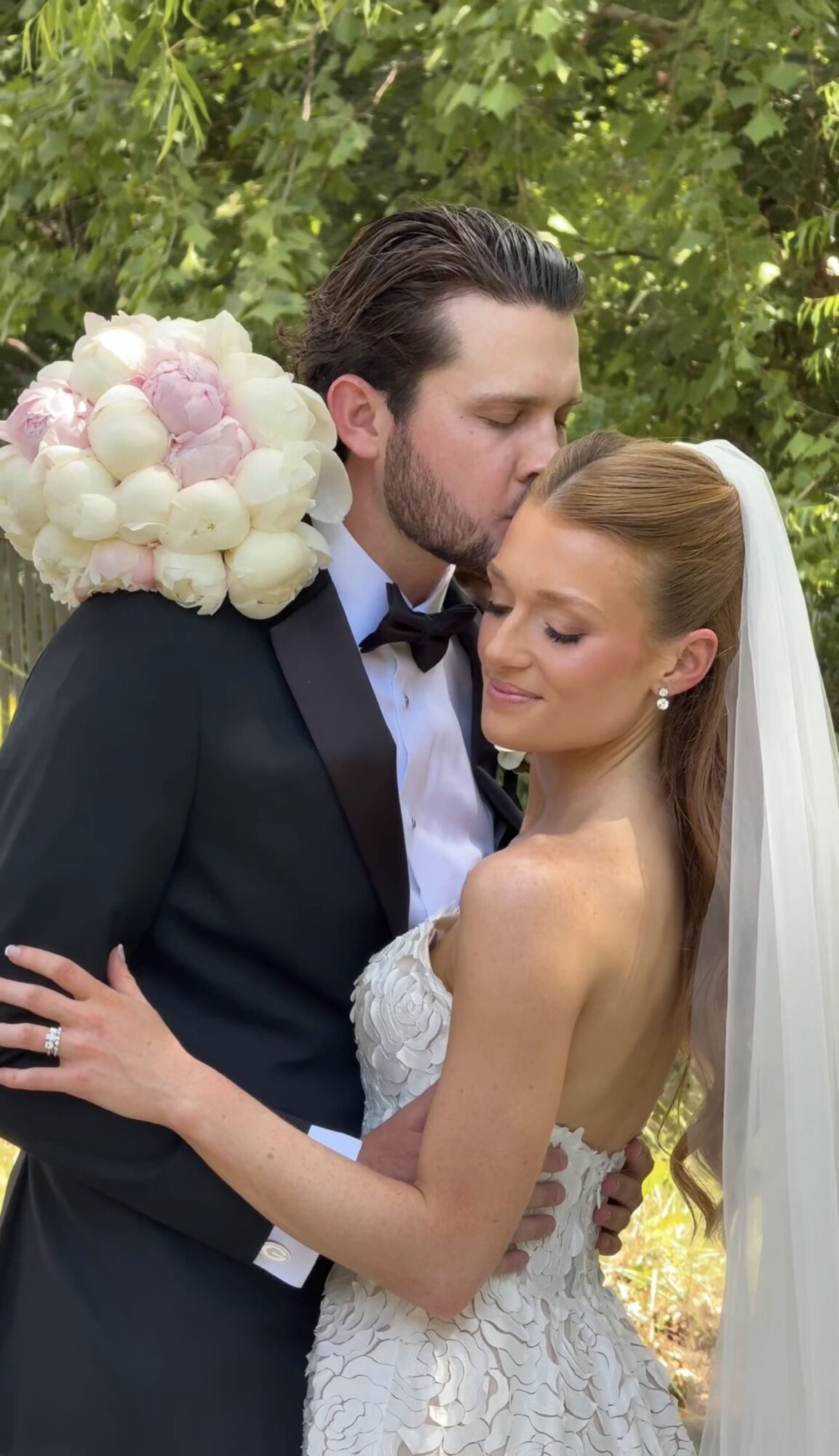 A bride and groom embrace outdoors, the groom kisses the bride's forehead, surrounded by green foliage.
