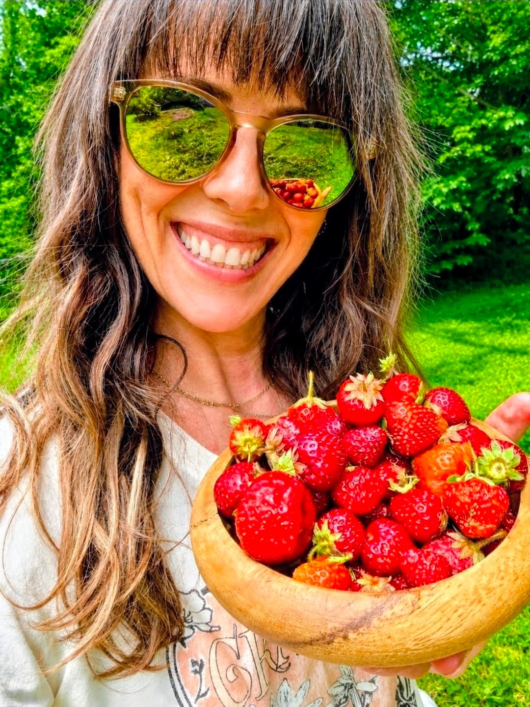 Woman with long hair and sunglasses smiling holding a bowl of strawberries outdoors in a green park.