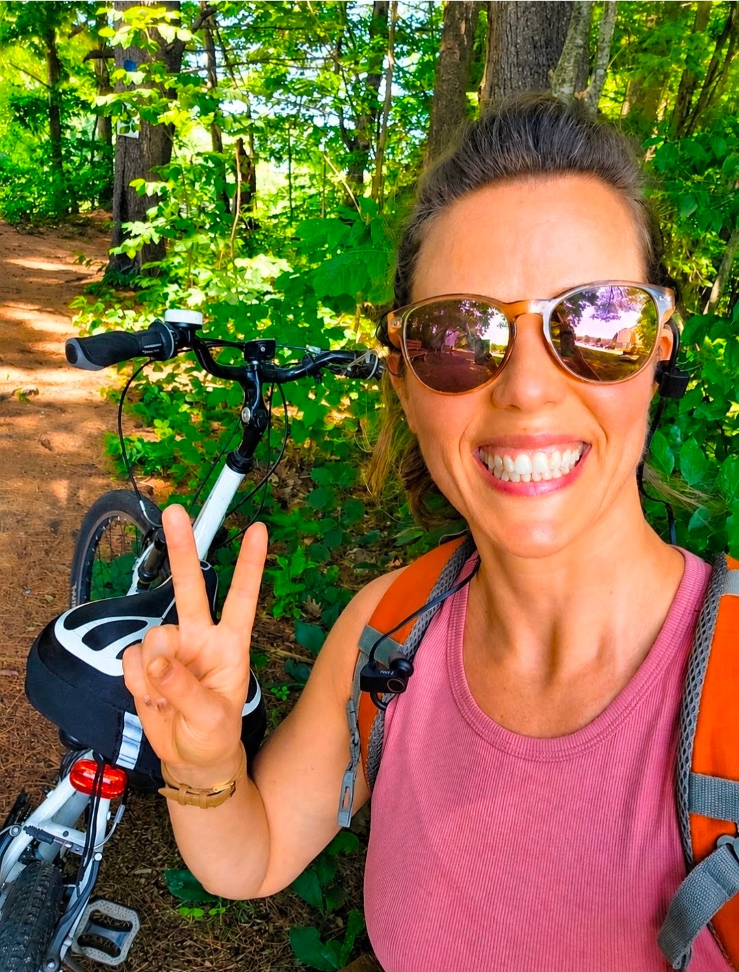 Woman smiling with sunglasses making a peace sign in a forested area with a bicycle behind her.