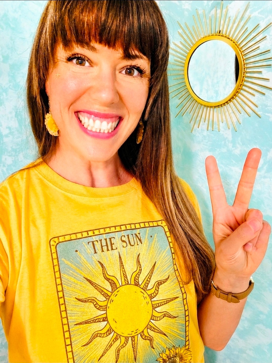 Smiling woman with brown hair and yellow shirt making a peace sign, standing in front of a sun-shaped mirror on a blue wall.