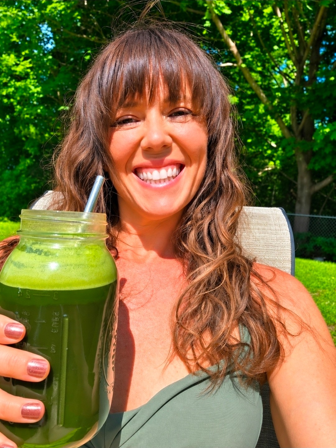 Smiling woman with long brown hair holding a green smoothie outdoors, trees in background.