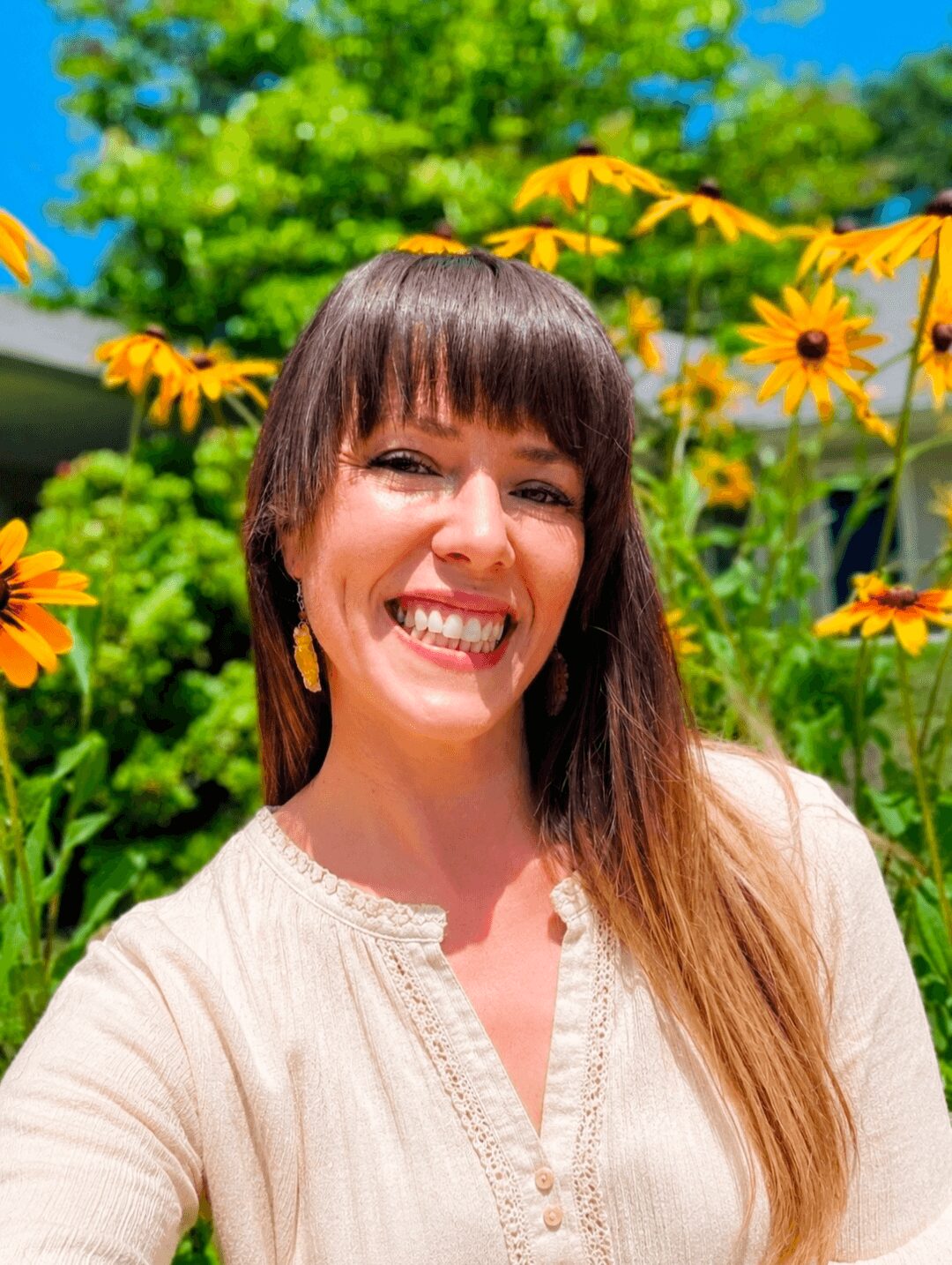 Smiling woman with long brown hair outdoors, surrounded by yellow flowers and green trees.