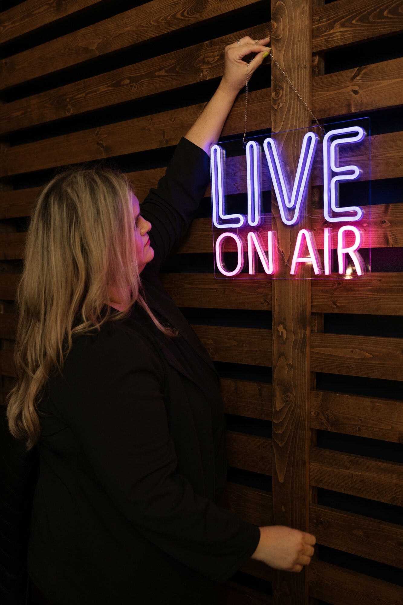 Woman with blonde hair reaching up to hang a neon sign that reads 'LIVE ON AIR' against a wooden wall.