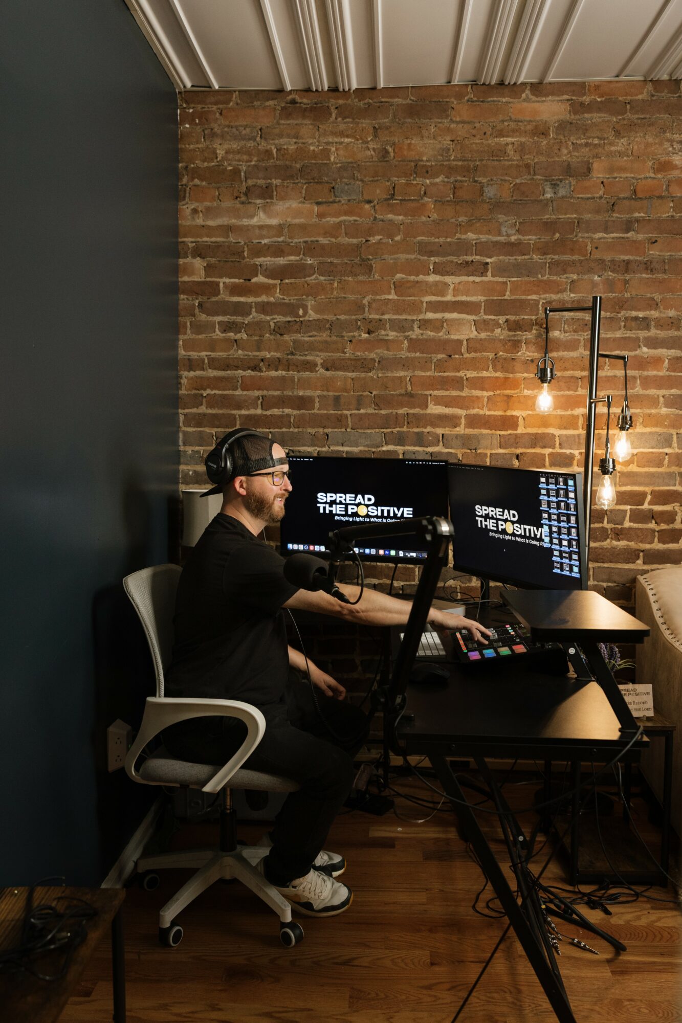 Person working at a desk with multiple monitors, brick wall background, hanging light bulbs, and wooden floor.