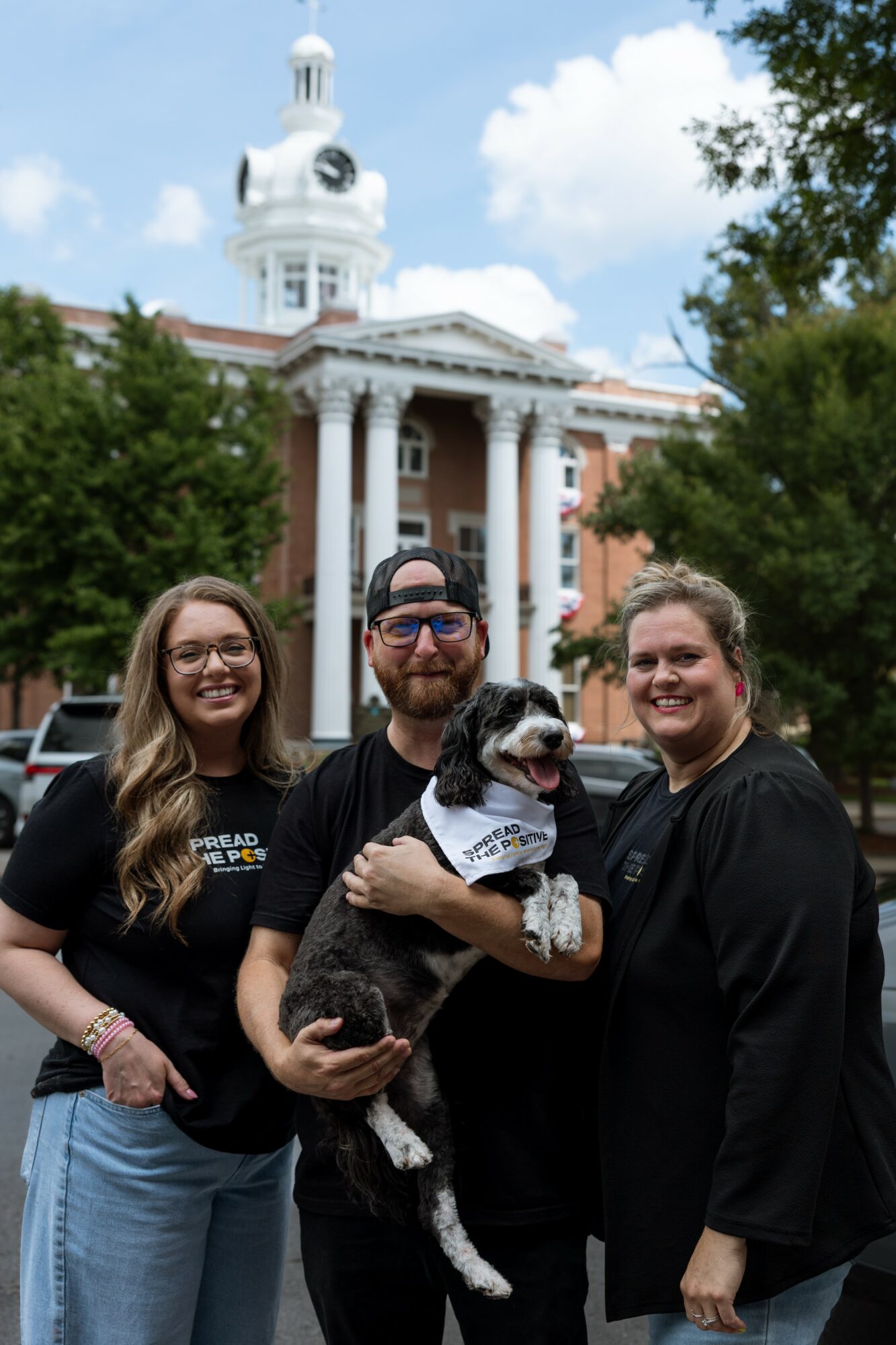 Three people and a dog stand in front of a historic building with a clock tower, trees, and a blue sky.