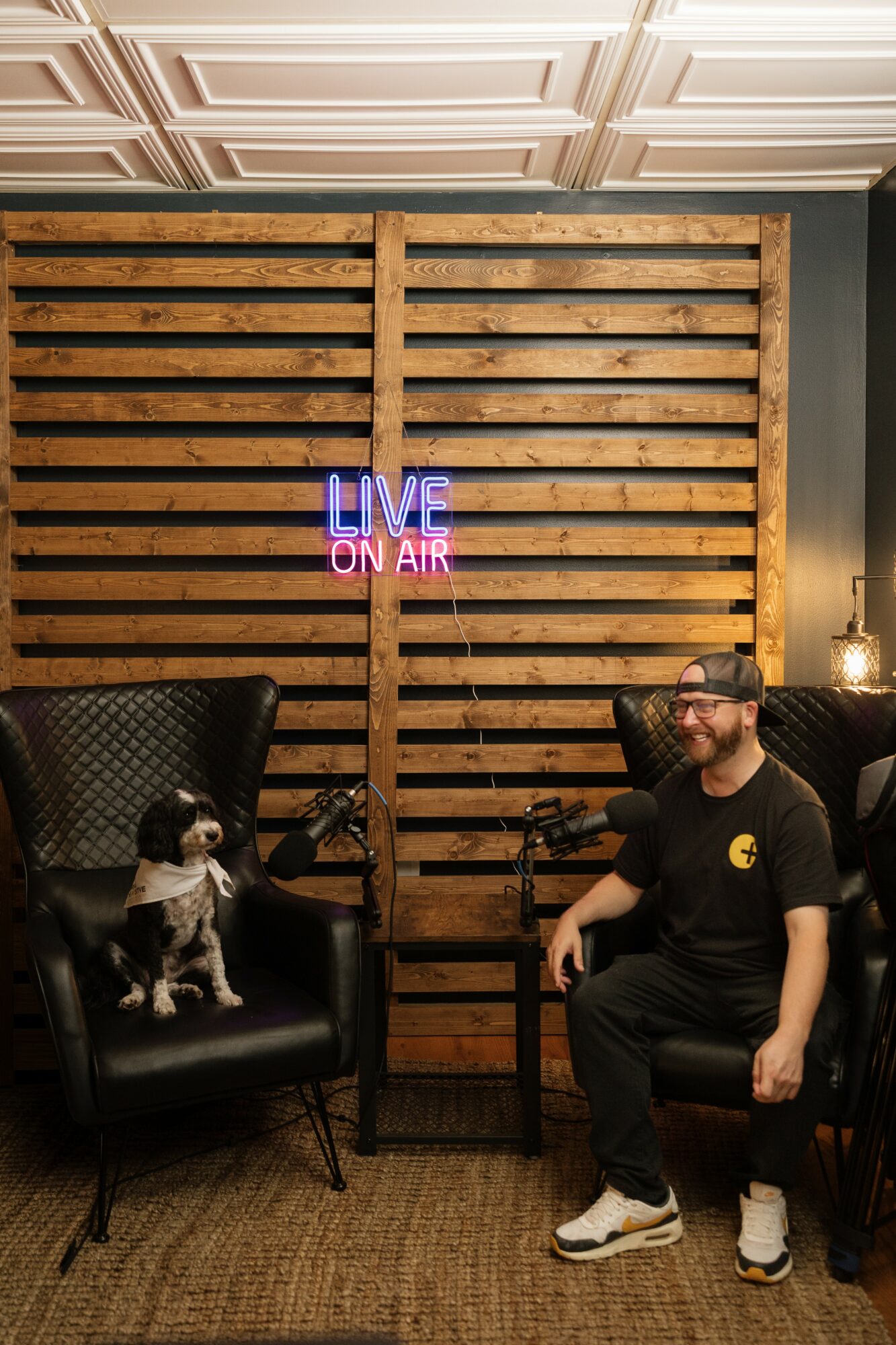 Man with glasses and beard sitting in a black chair, dog on another chair, wooden slat wall with neon sign reading 'LIVE ON AIR', lamp, and textured rug.