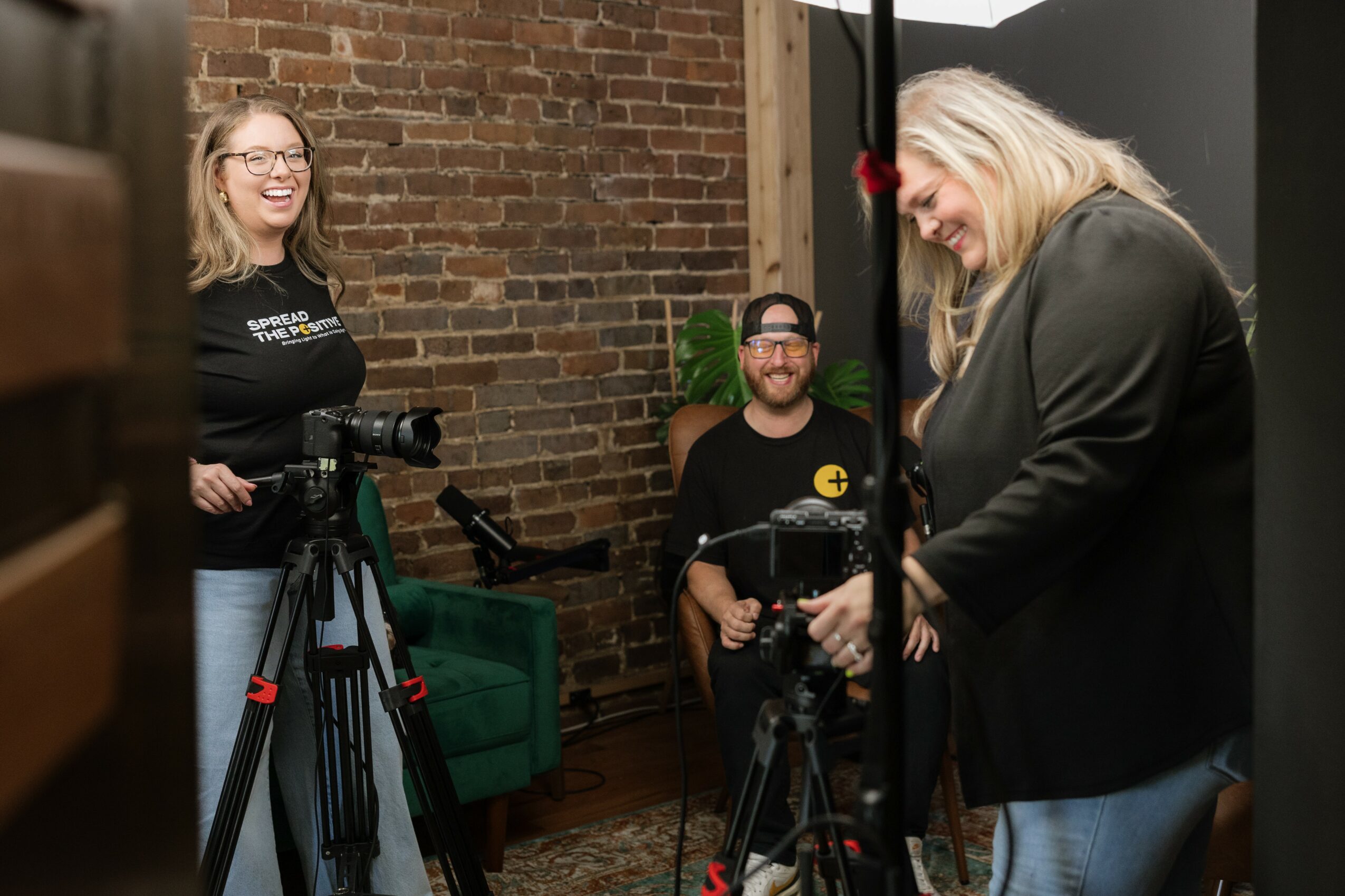 Three people in a room with a brick wall, two women and one man, preparing filming equipment.