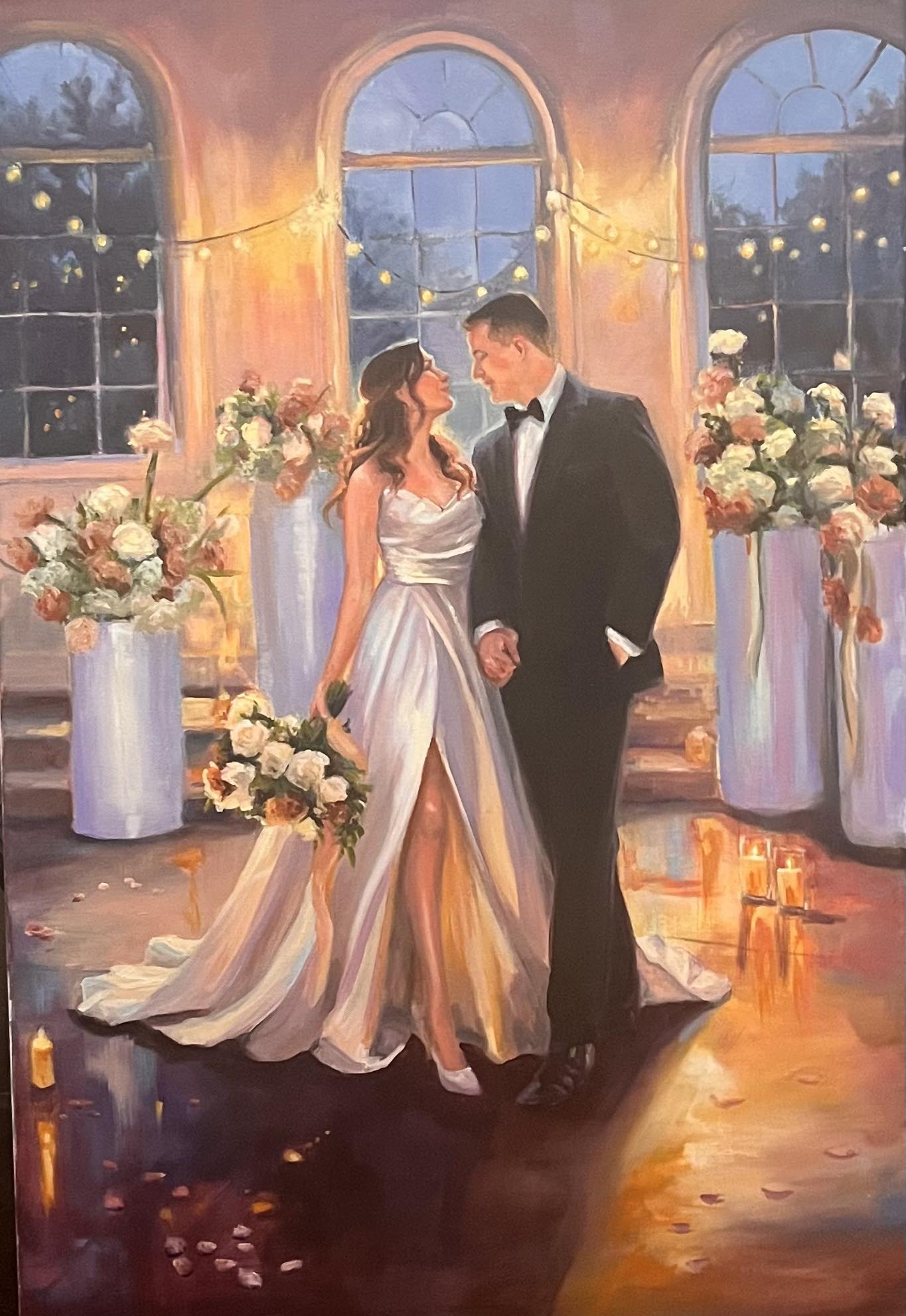 A bride and groom holding hands in a decorated indoor setting with large windows, flowers, and candles, during evening or night.