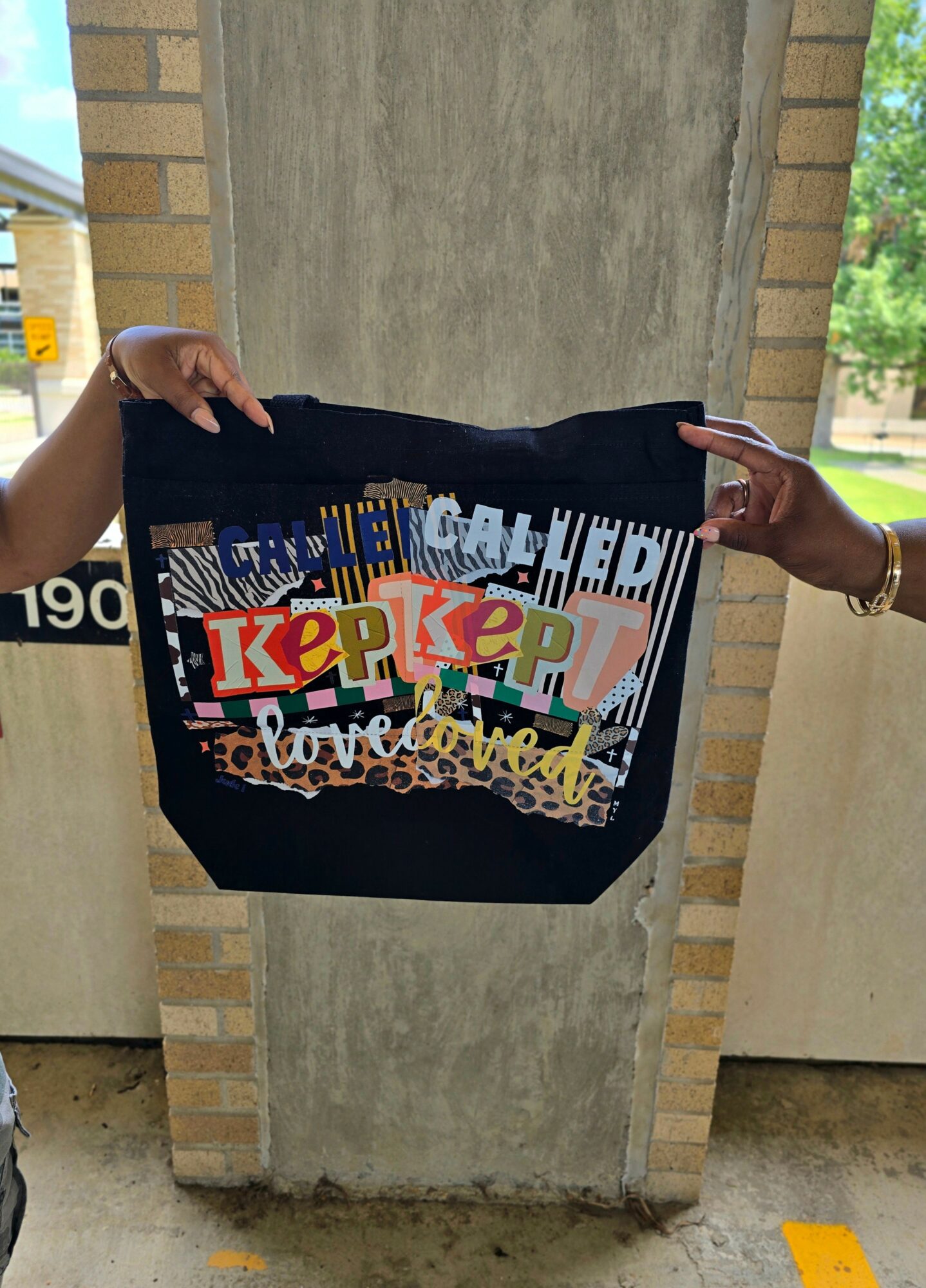 Two people hold a black bag with colorful cut-out letters and designs, standing outdoors near a brick and concrete structure.