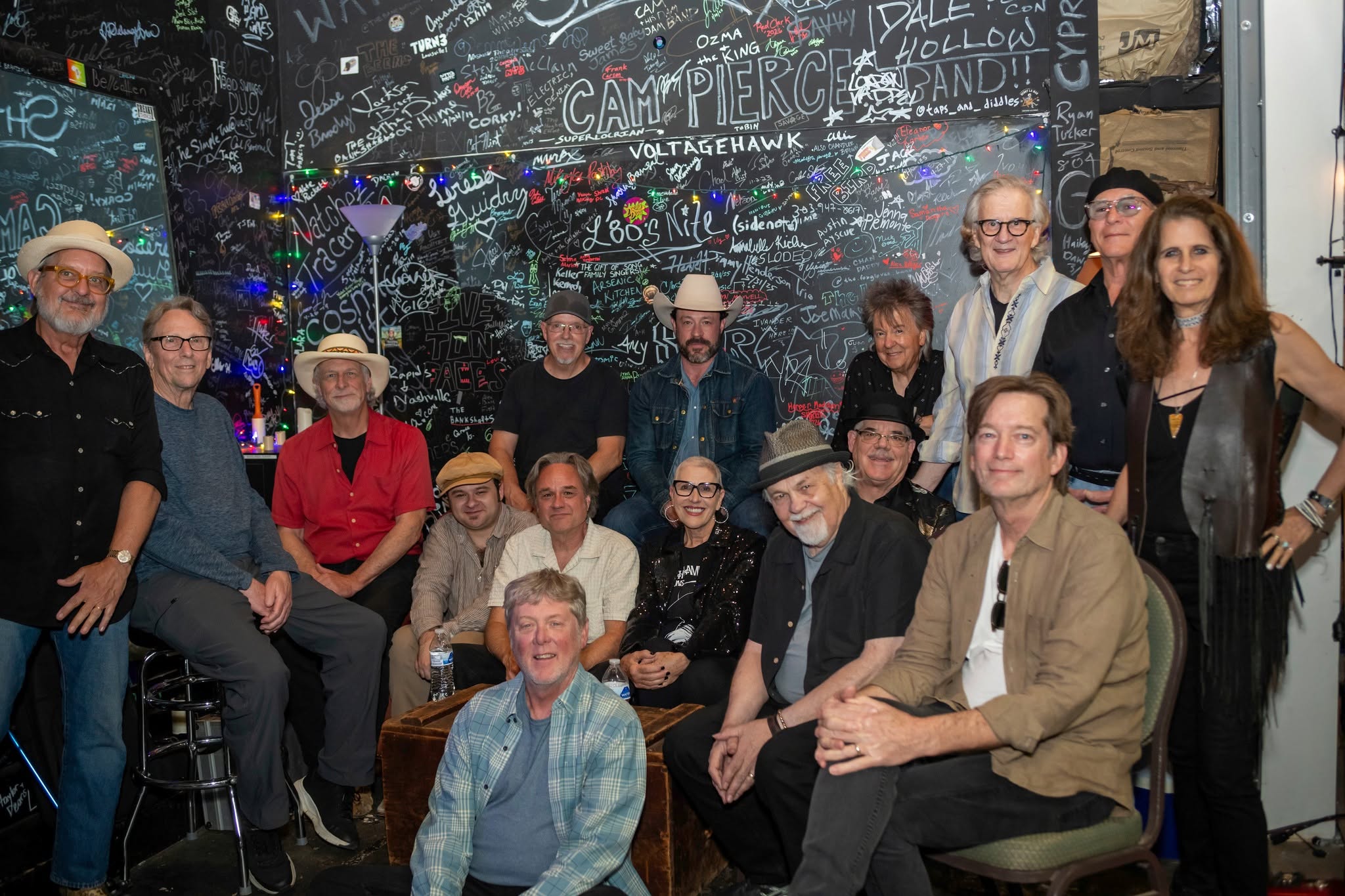 Group of people gathered indoors, some sitting and some standing, in front of a decorated wall with colorful writing and lights.