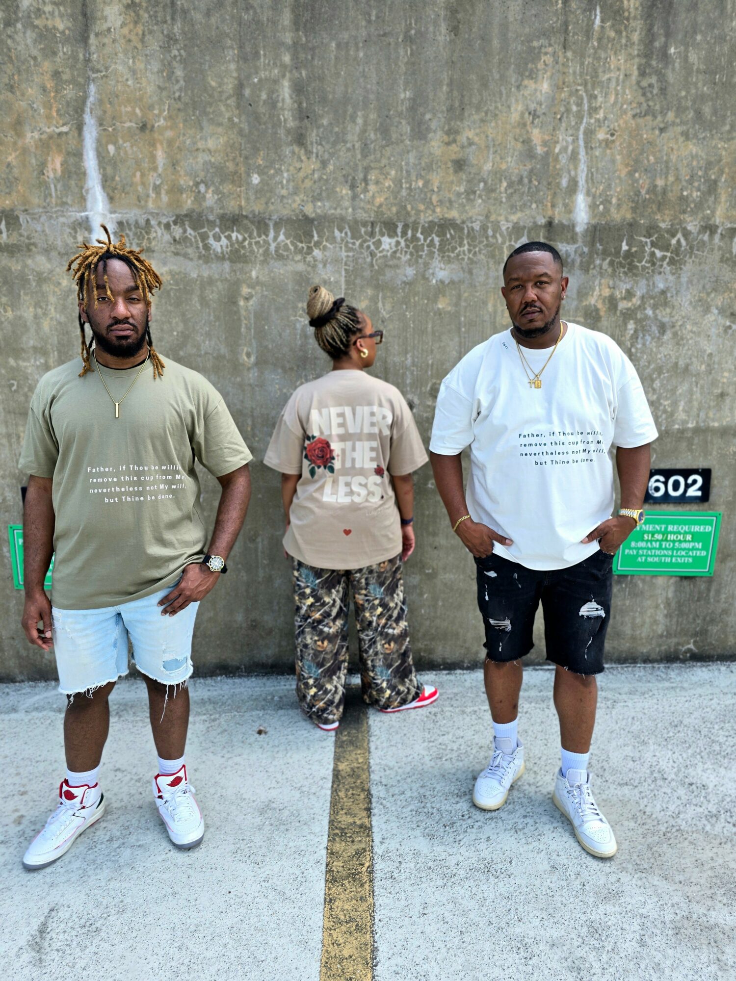 Three people standing against a concrete wall, two men on sides and a woman in the middle, facing different directions.