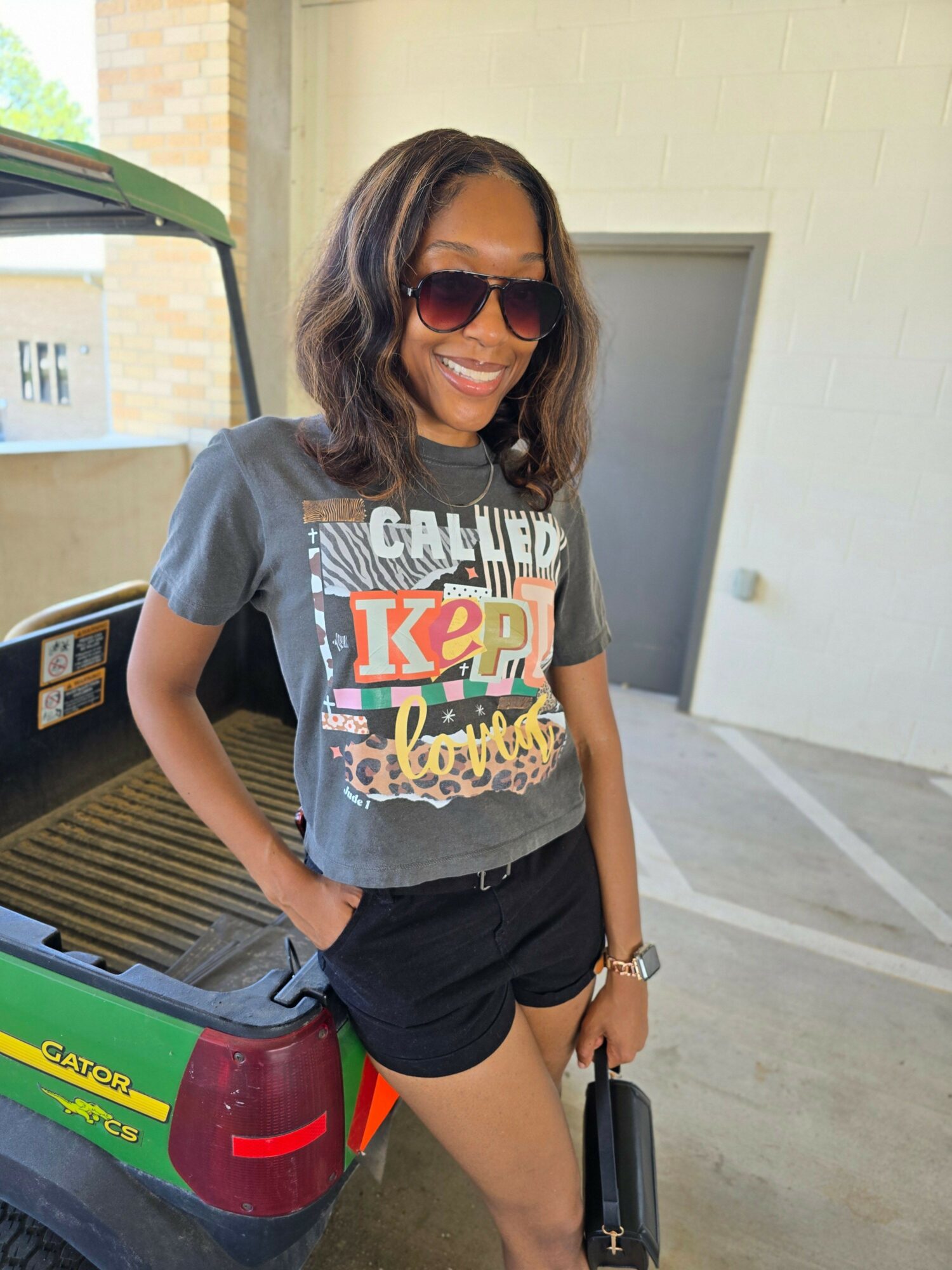 Woman with sunglasses wearing a graphic T-shirt and shorts, standing outdoors next to a colorful object, smiling.