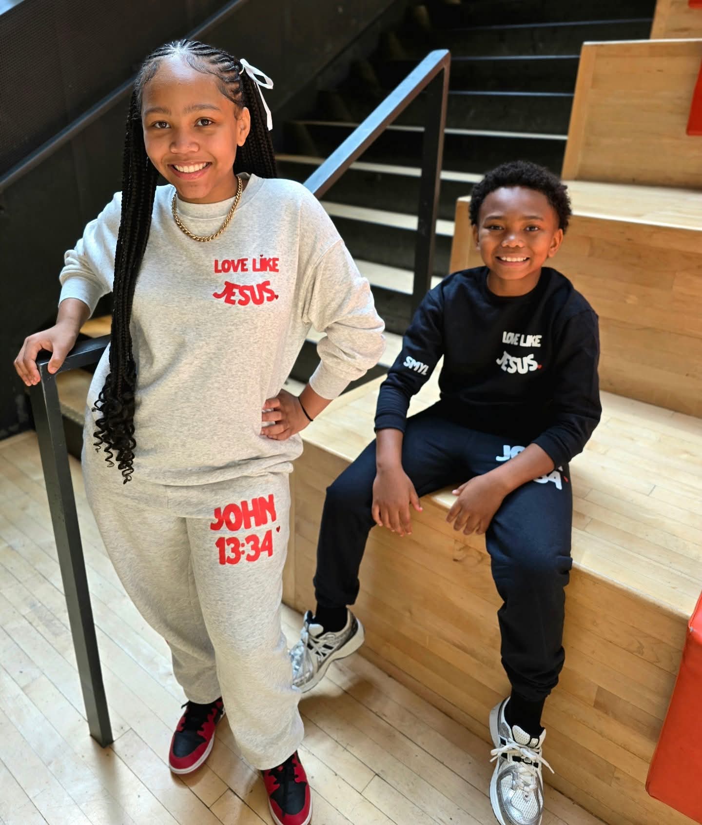 Two children sitting on wooden stairs, smiling, one girl with long braided hair and a boy with short curly hair, both wearing casual clothes with religious messages.