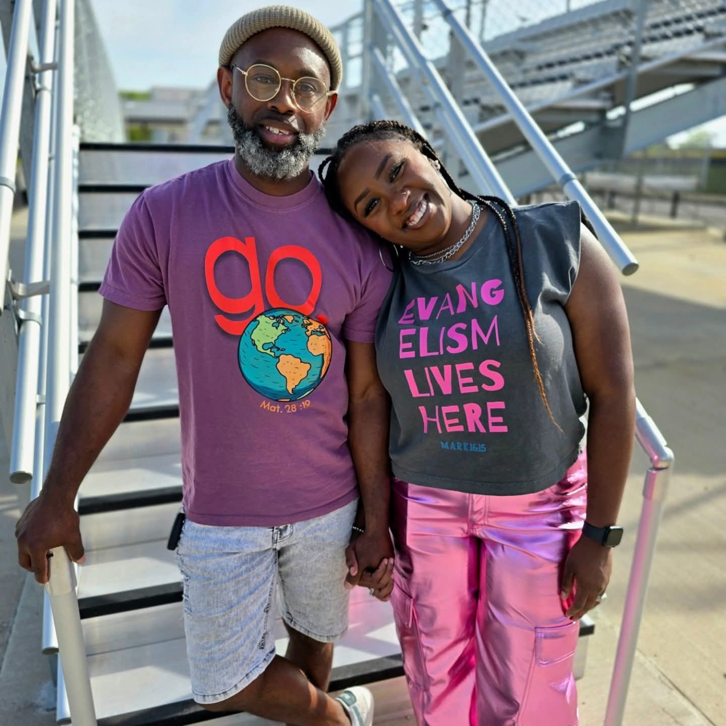 A smiling man and woman holding hands on outdoor stairs, both wearing casual clothing and glasses.