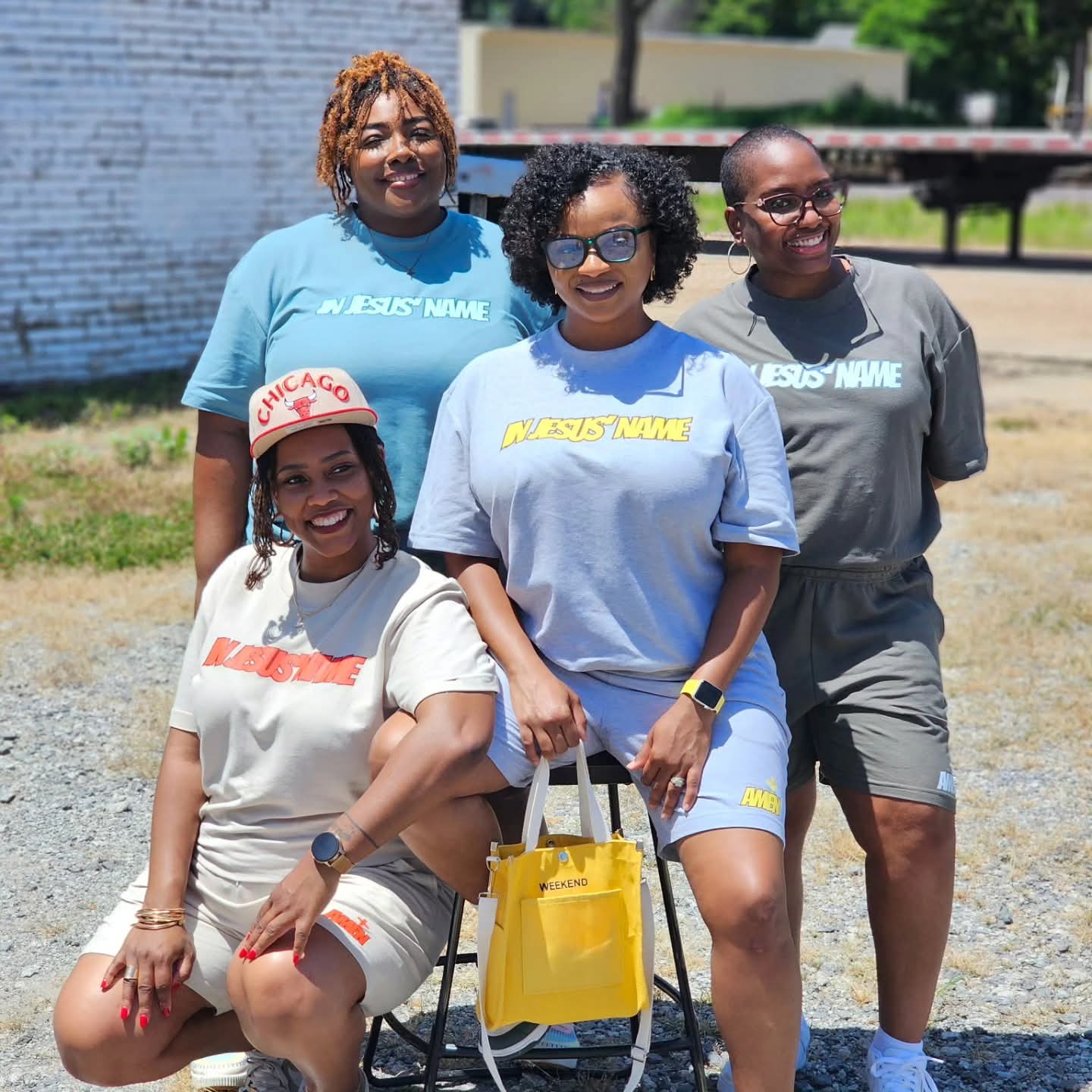 Four women outdoors, smiling, wearing casual clothes and sunglasses, with a yellow bag, in front of a white brick wall.