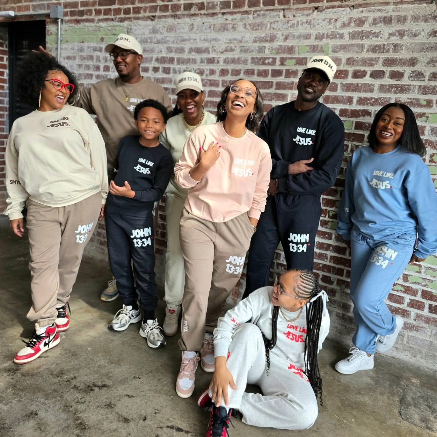 Group of eight diverse people smiling and laughing against a brick wall, some wearing matching hats and sweatshirts.