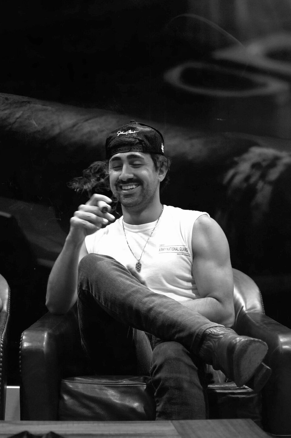 Man smiling, wearing a cap backwards, sitting with legs crossed, holding a cigarette, in a dark setting.