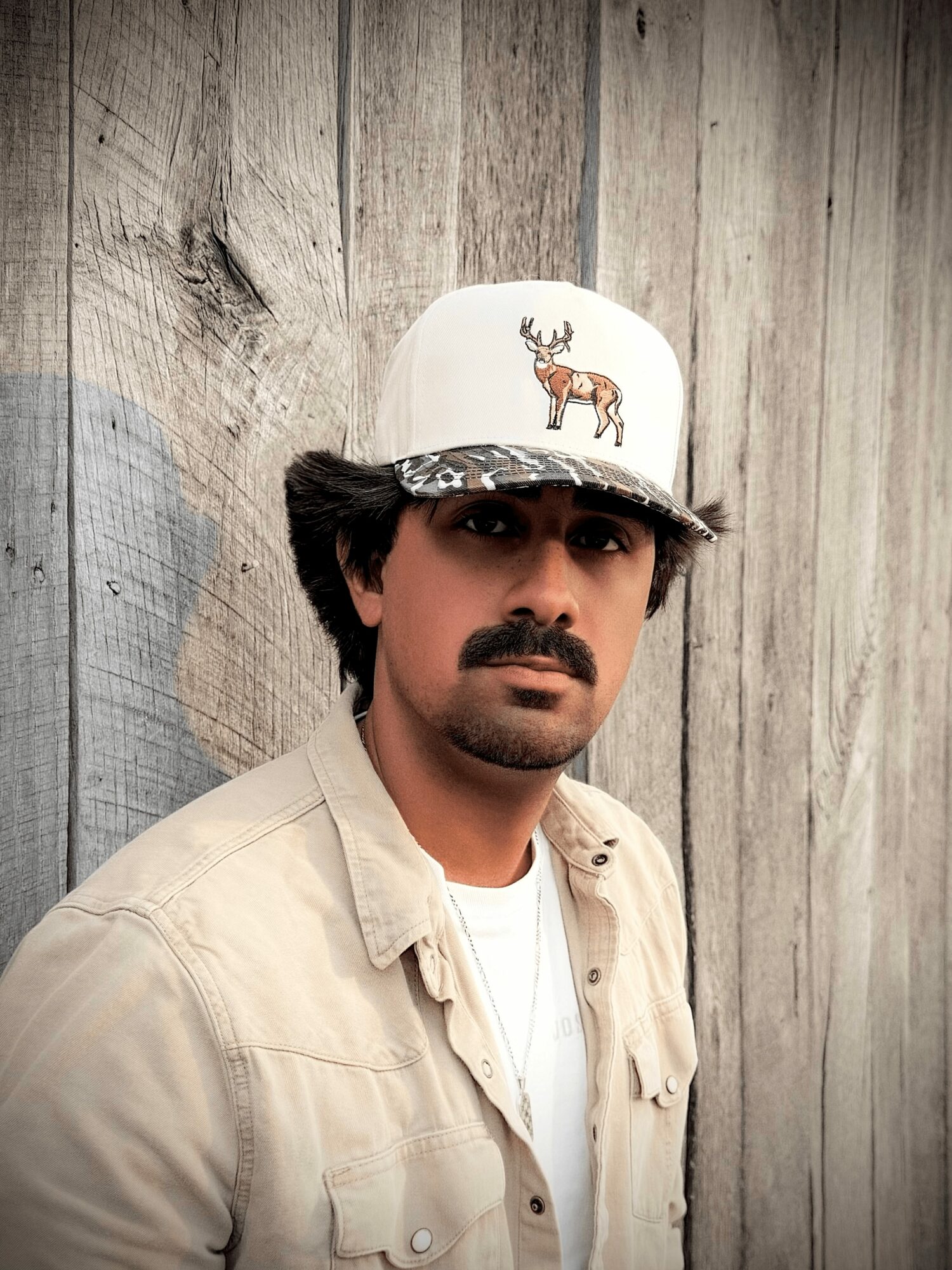 Man with a mustache and dark hair wearing a white cap with a moose and deer design, beige shirt, standing against wooden background.