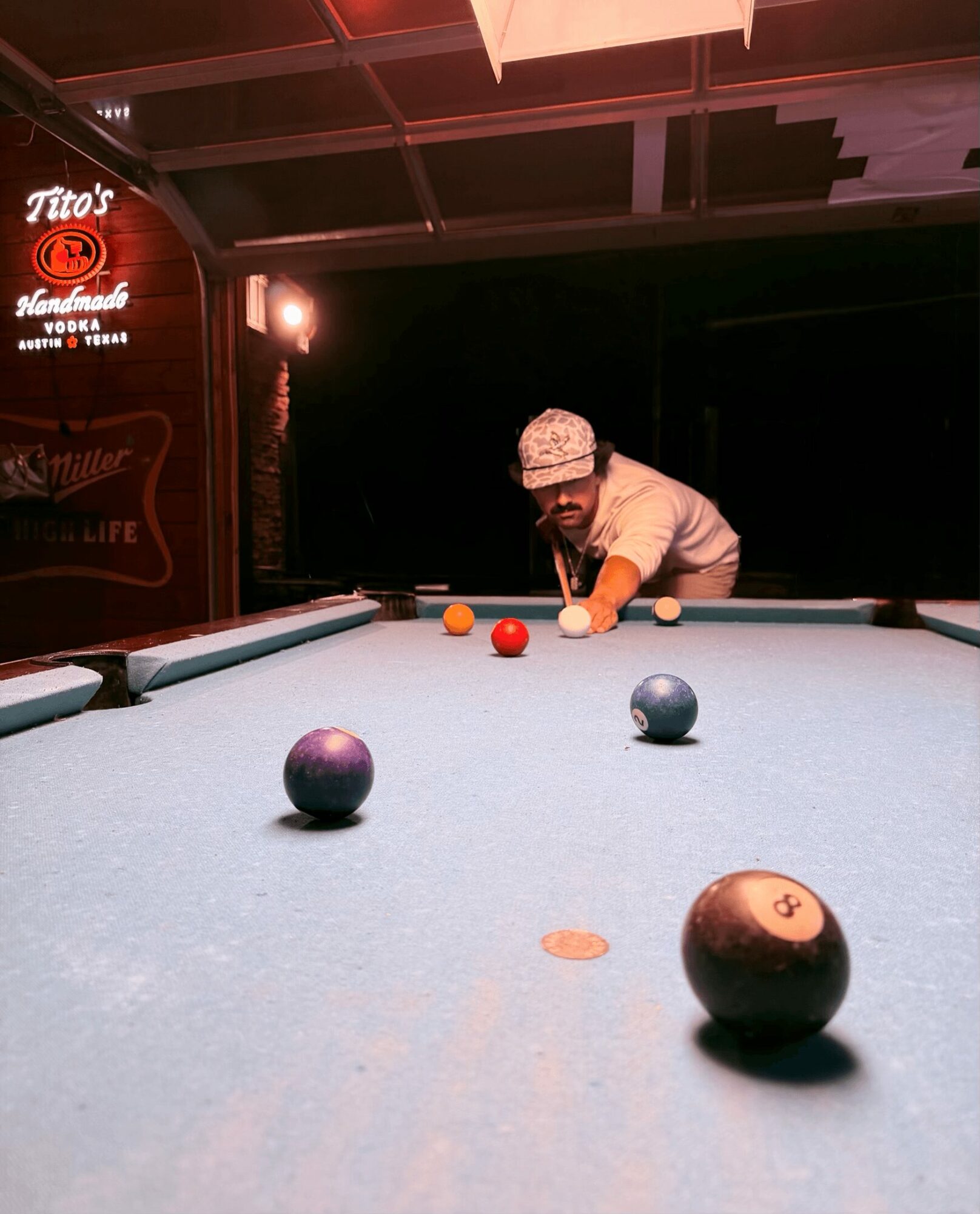 Person playing pool at a bar, aiming at balls on the table, with neon signs in the background.