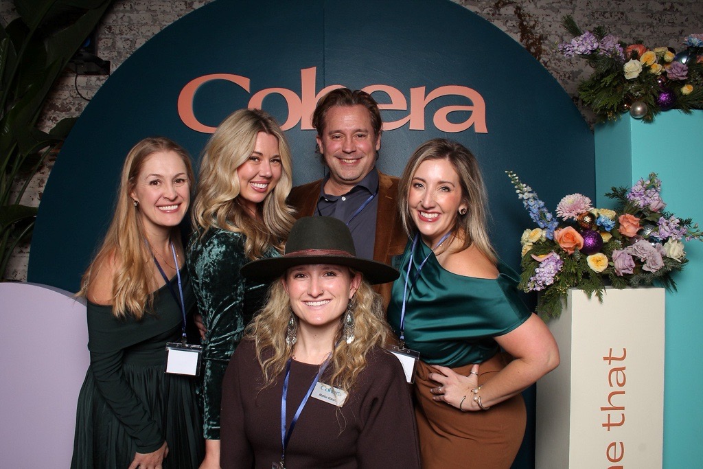 Five women and one man pose together in front of a backdrop with the word 'Colore' and floral decorations, smiling.