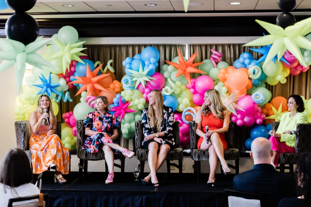Five women sit on a stage with colorful balloon decorations behind them, speaking into microphones and engaging with the audience.