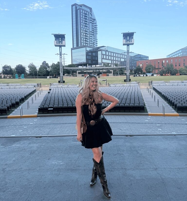 Young woman in black dress and boots standing outdoors with city buildings in background.