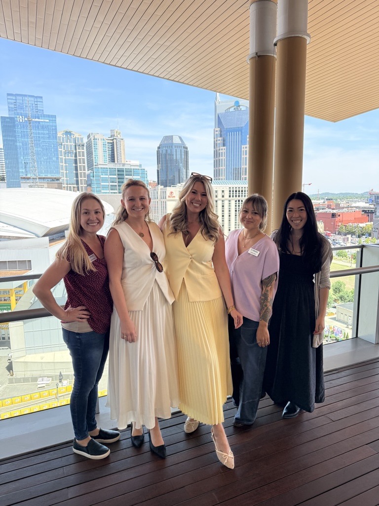 Five women standing on a balcony with city skyline in background, smiling, dressed in casual and semi-formal clothing.
