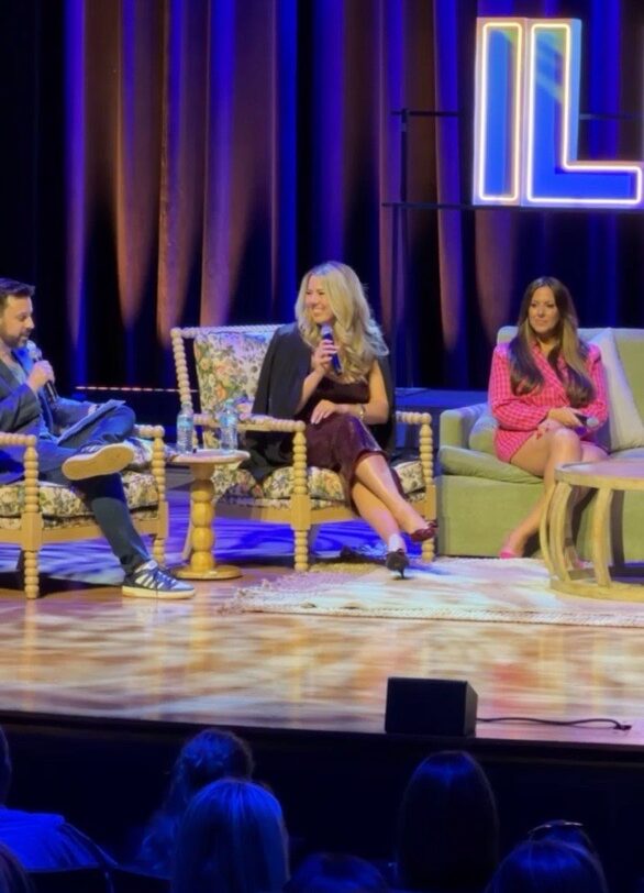 Three people sit on stage with colorful curtains and a neon sign in the background, engaged in conversation.