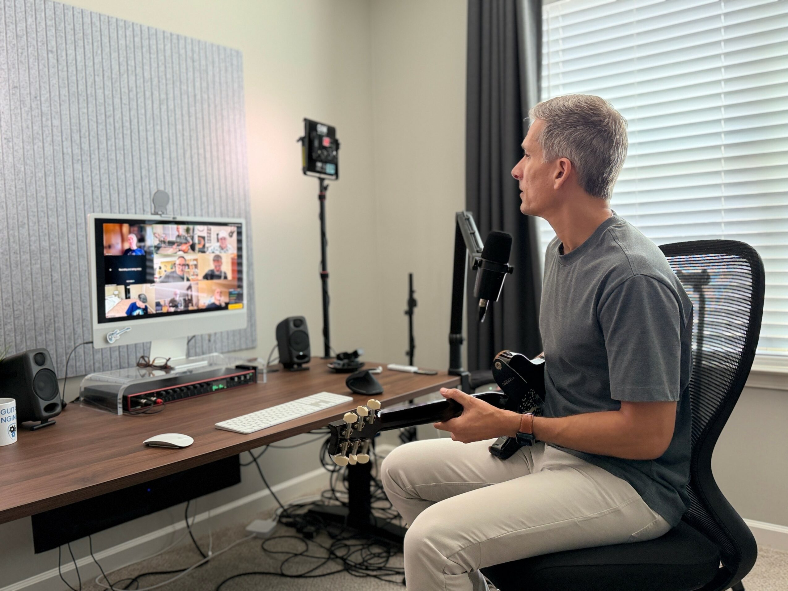 Man sitting in a chair in front of a computer monitor during a video call, with a microphone and camera setup nearby.