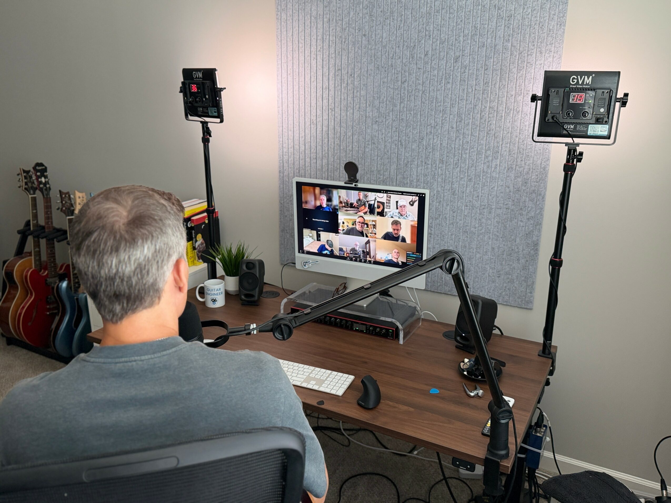 Person with gray hair sitting at desk in front of computer monitor with multiple video call participants, two lights on stands, and guitars in corner.