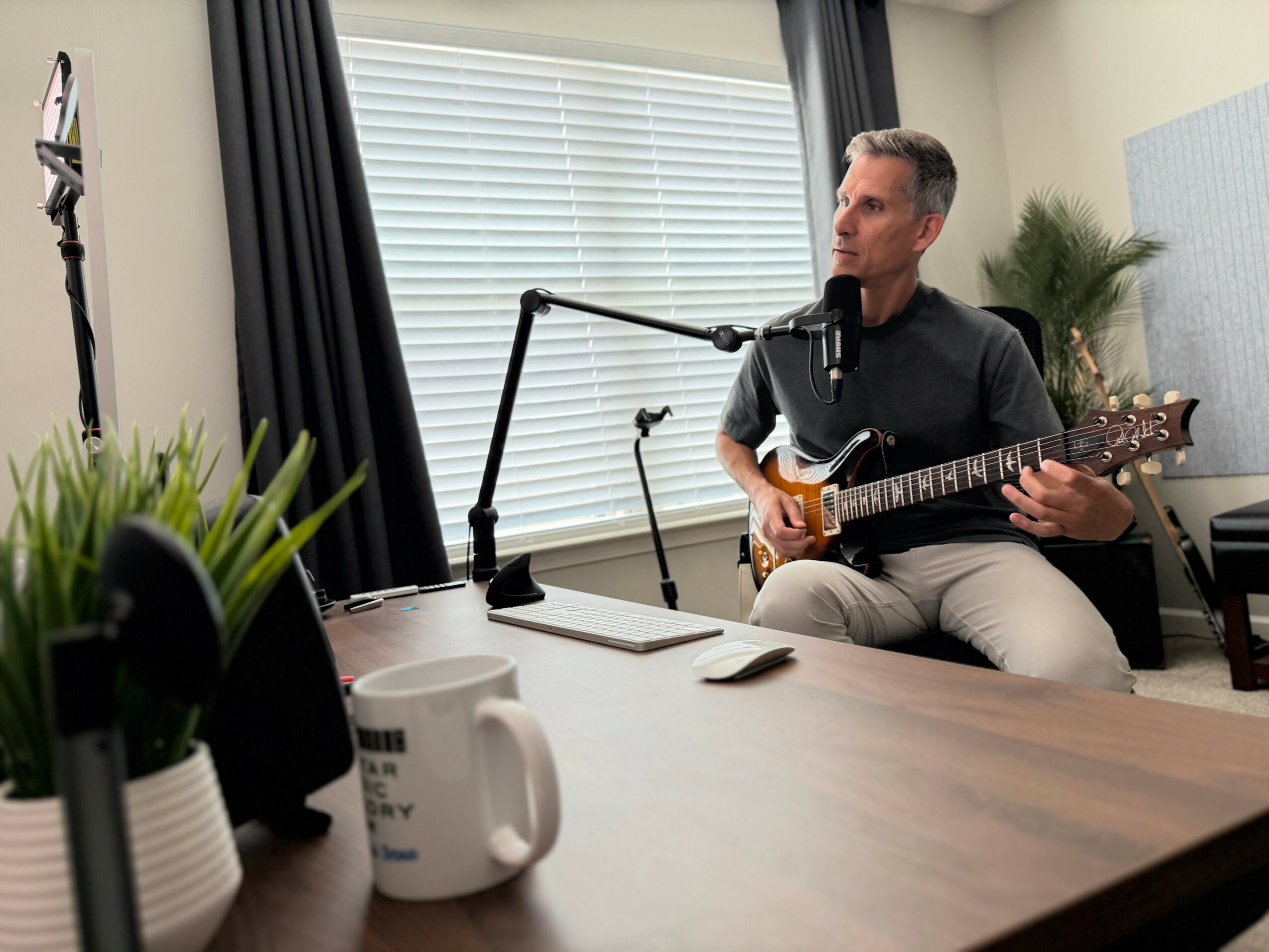 Man playing guitar in a room with window blinds, plants, and a microphone, sitting on a chair, with a table in front.