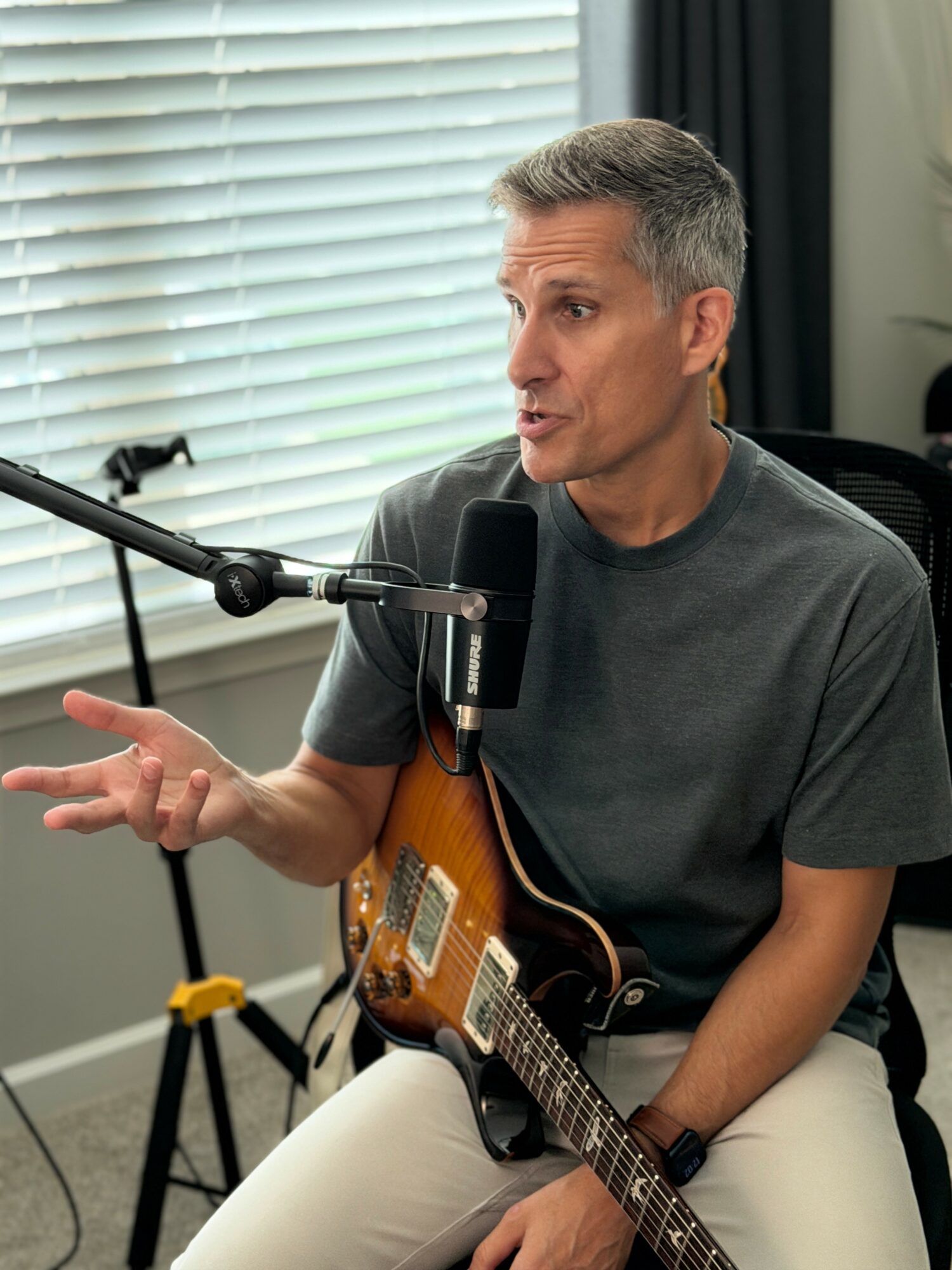 Man speaking into microphone while holding an electric guitar, gesturing with his hand, in a room with window blinds.