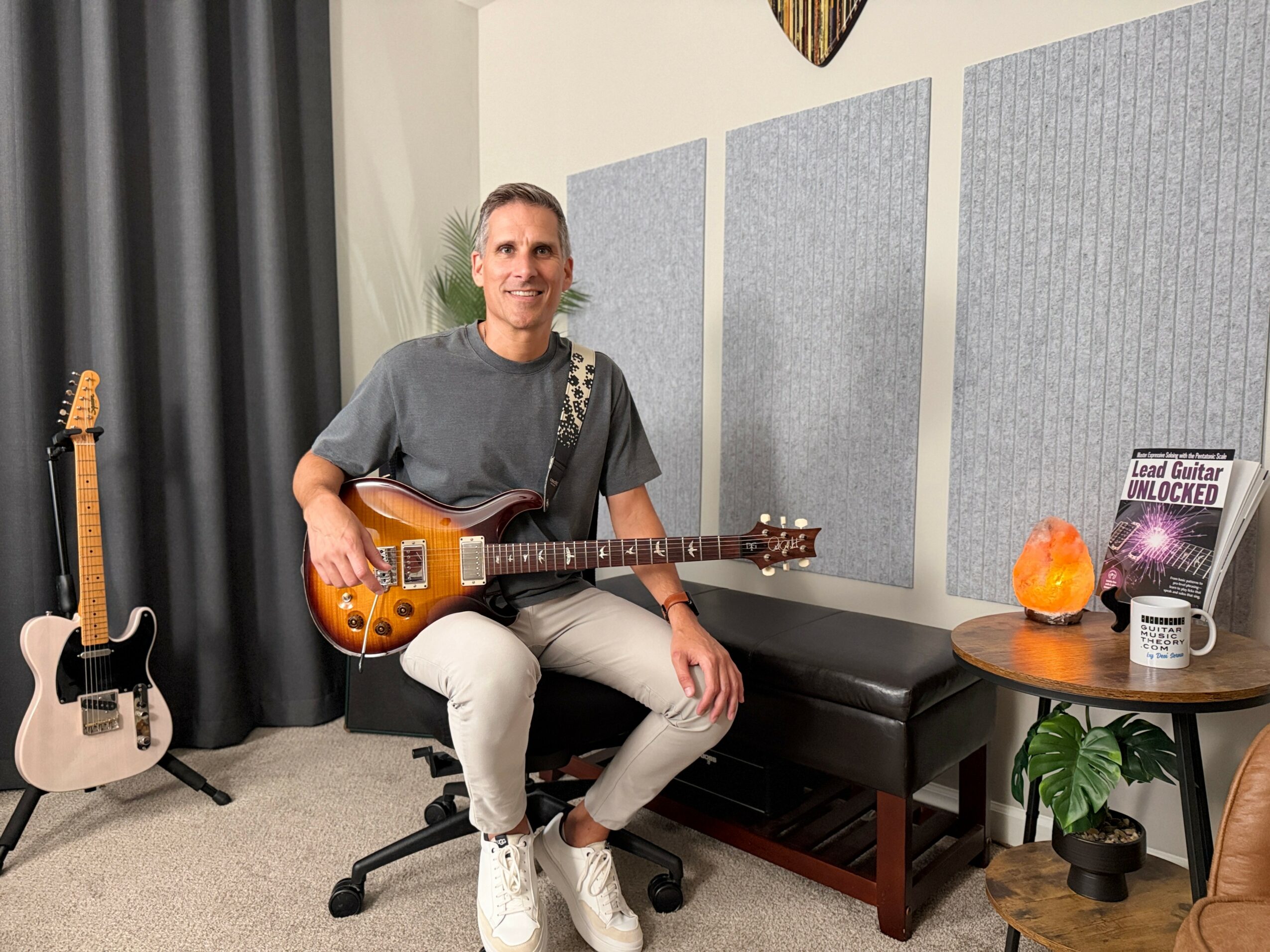 Person sitting on a chair holding an electric guitar in a music room, with another guitar on a stand nearby and a table with a salt lamp and plant.